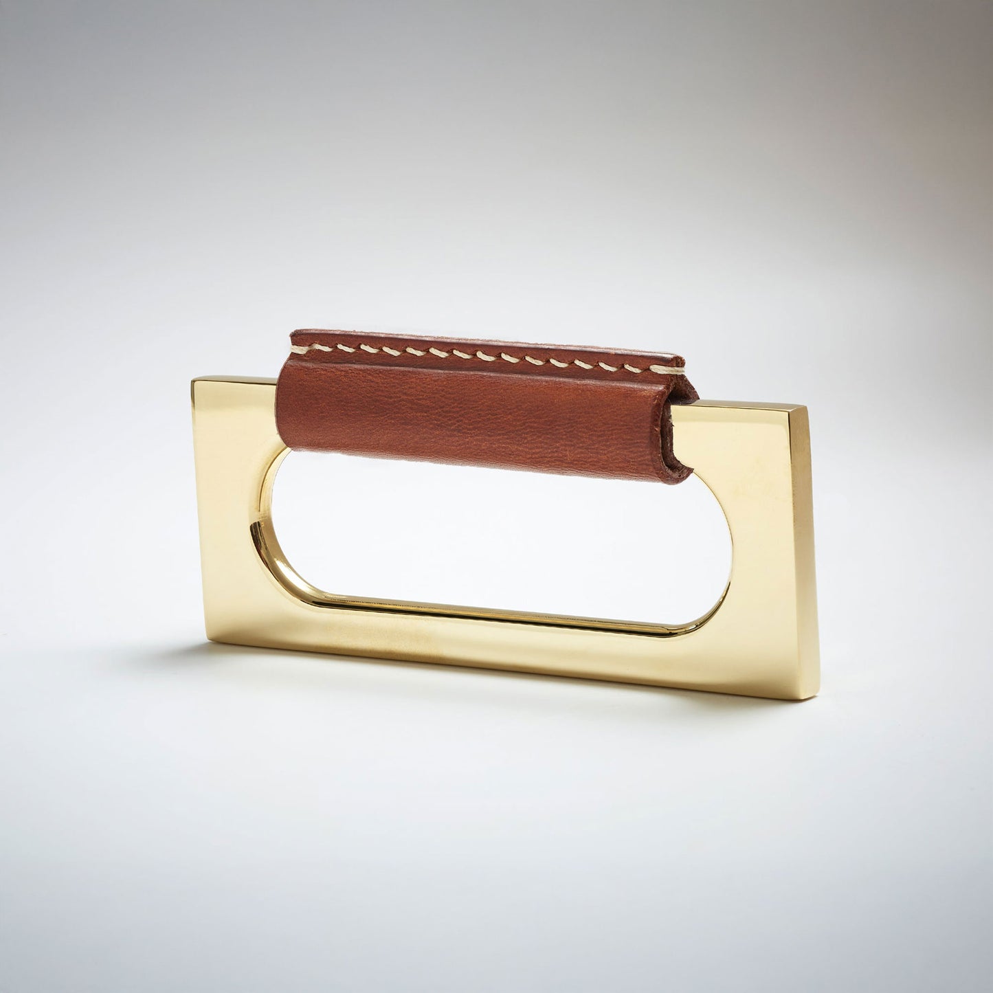 A modern rectangular metal cabinet pull with a central cutout, featuring a light brown leather strap stitched across the top, shown on a clean grey studio background.