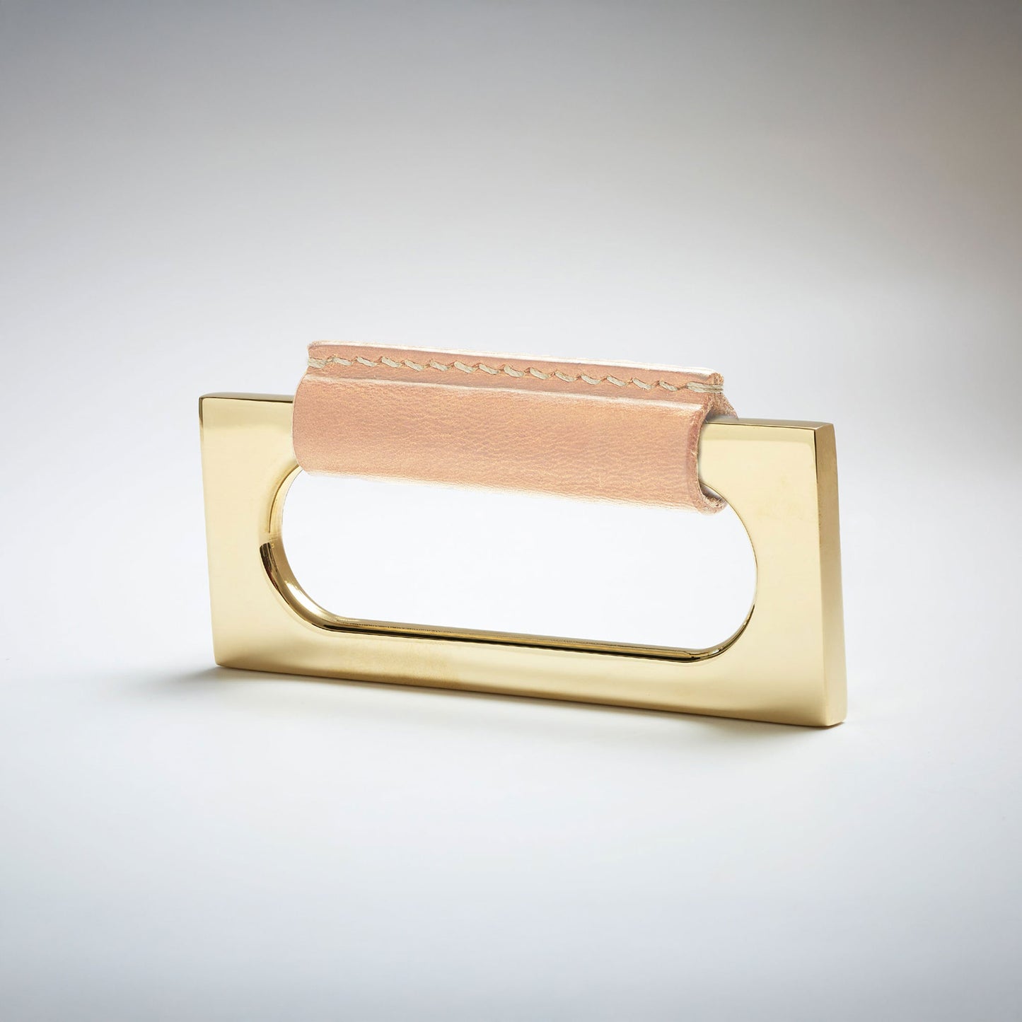 A modern rectangular metal cabinet pull with a central cutout, featuring a light tan leather strap stitched across the top, shown on a clean grey studio background.