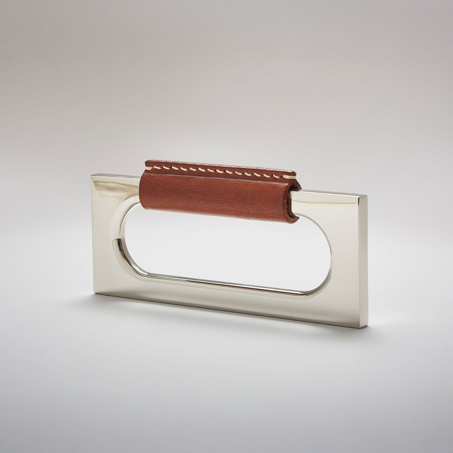 A modern rectangular metal cabinet pull with a central cutout, featuring a light brown leather strap stitched across the top, shown on a clean grey studio background.