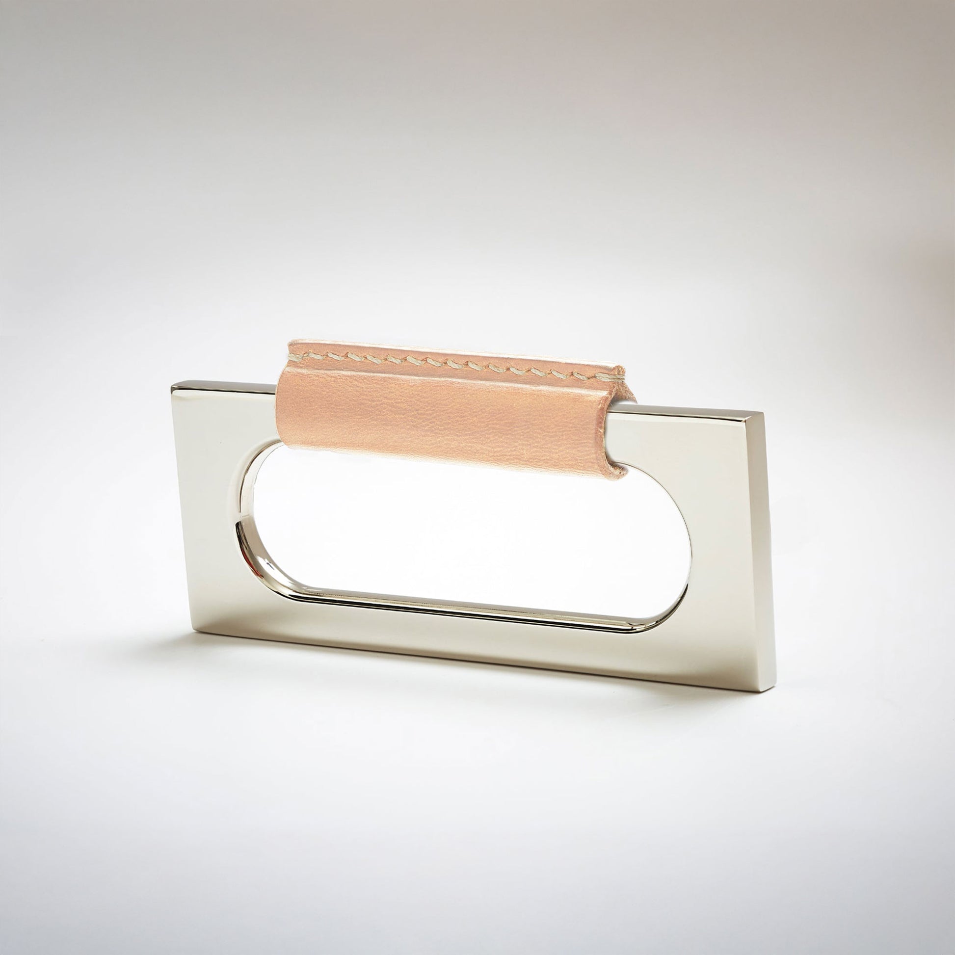 A modern rectangular metal cabinet pull with a central cutout, featuring a light tan leather strap stitched across the top, shown on a clean grey studio background.