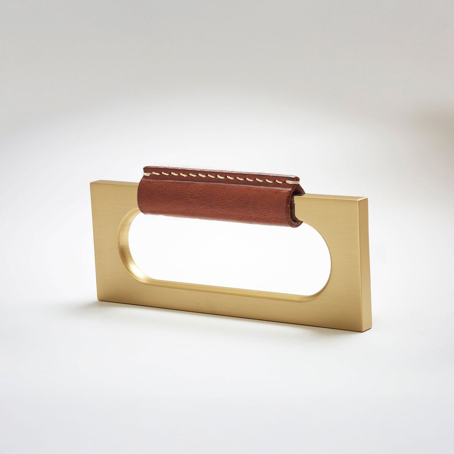 A modern rectangular metal cabinet pull with a central cutout, featuring a light brown leather strap stitched across the top, shown on a clean grey studio background.