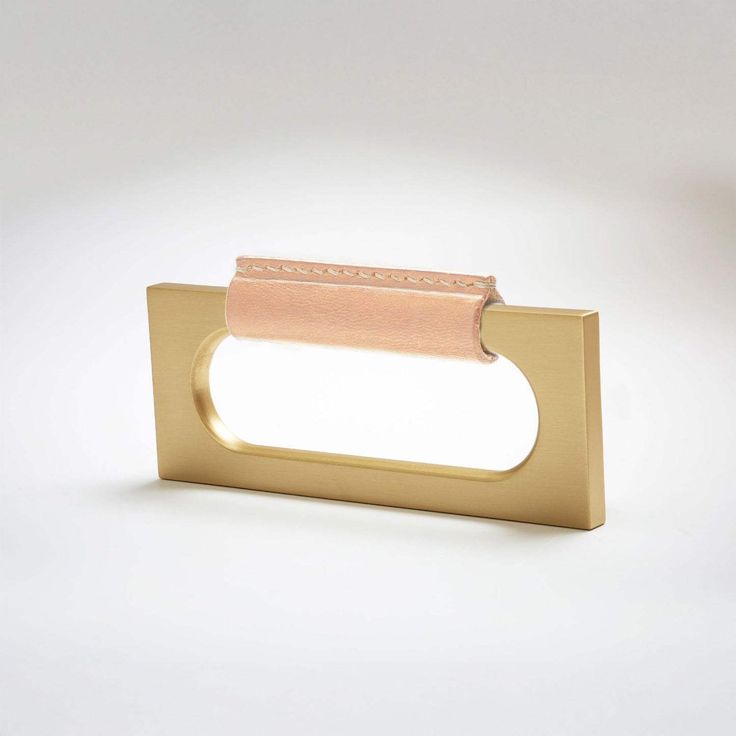 A modern rectangular metal cabinet pull with a central cutout, featuring a light tan leather strap stitched across the top, shown on a clean grey studio background.