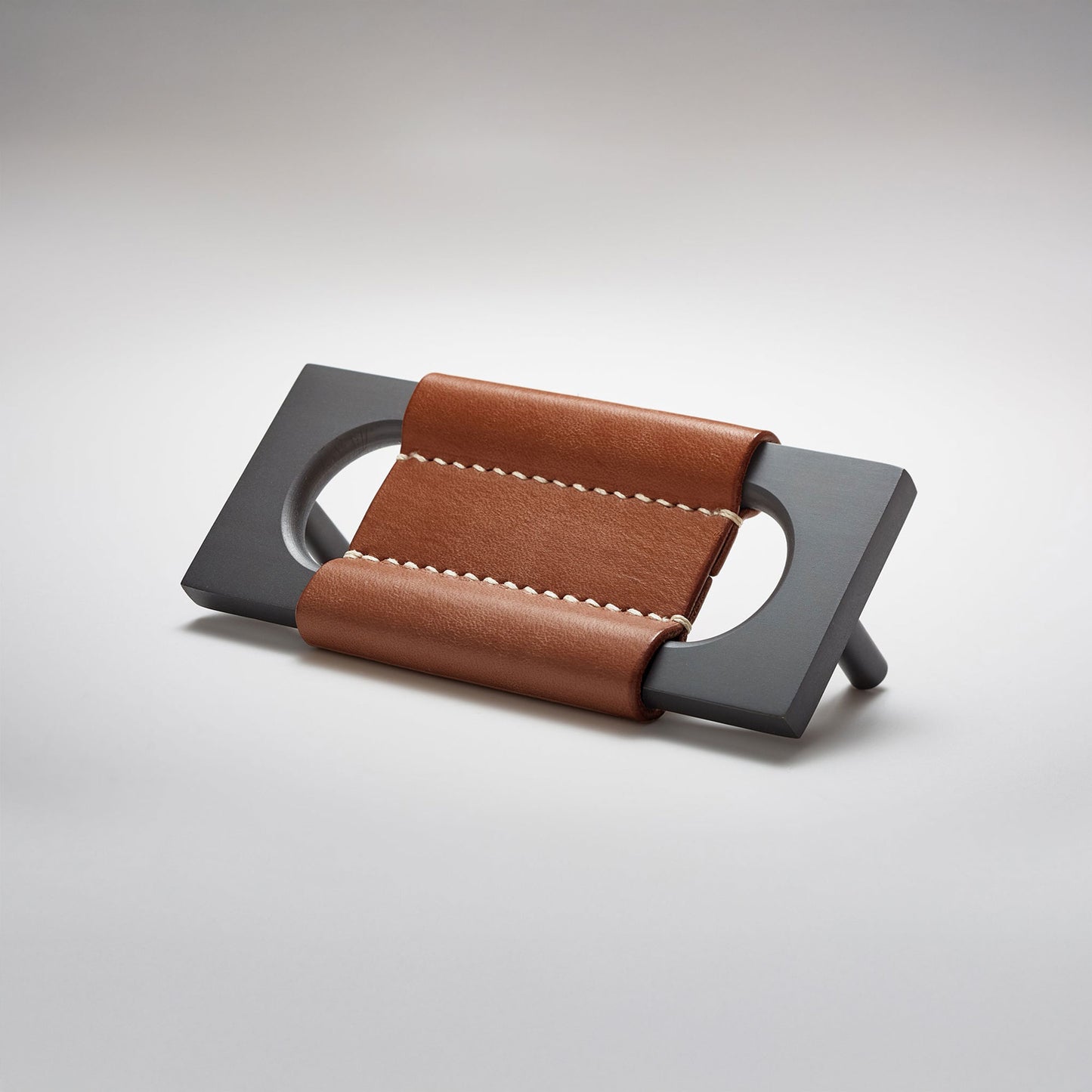 A modern rectangular metal cabinet pull with a central cutout, featuring a light brown leather strap stitched across the top, shown on a clean grey studio background.