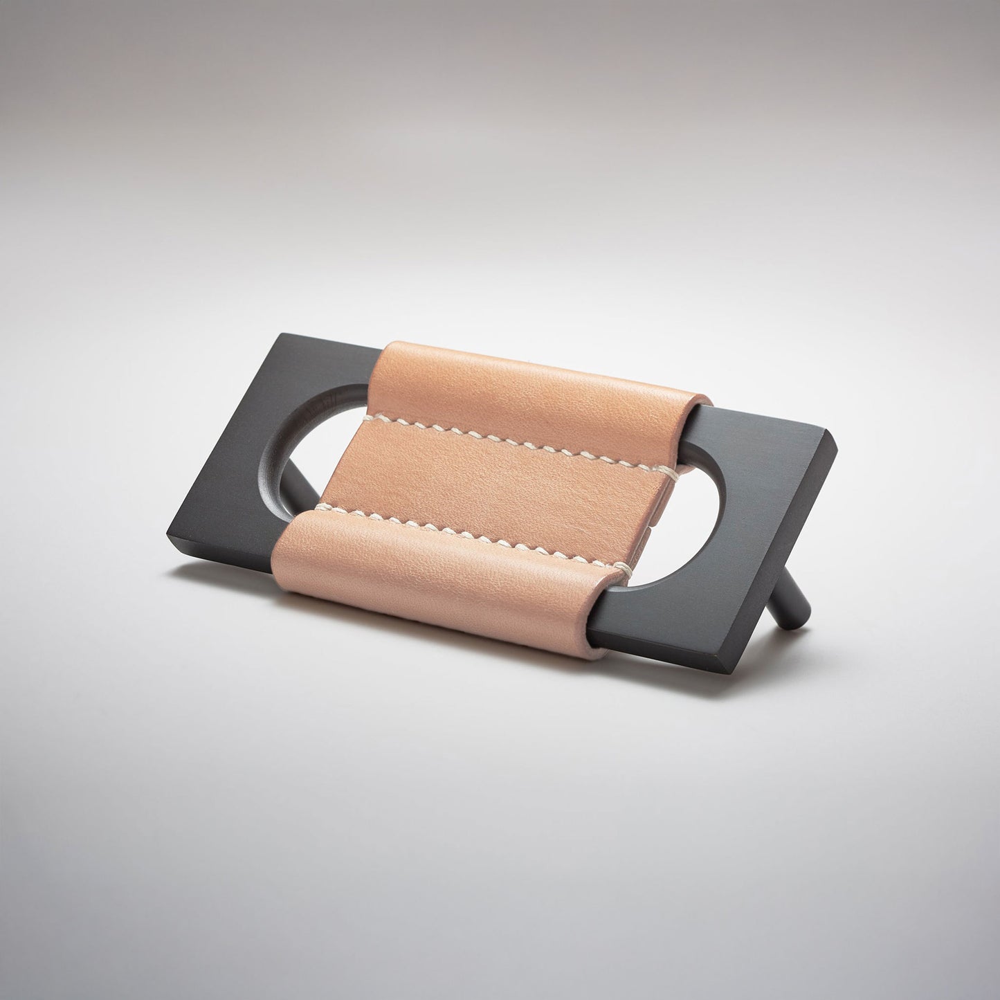 A modern rectangular metal cabinet pull with a central cutout, featuring a light tan leather strap stitched across the top, shown on a clean grey studio background.