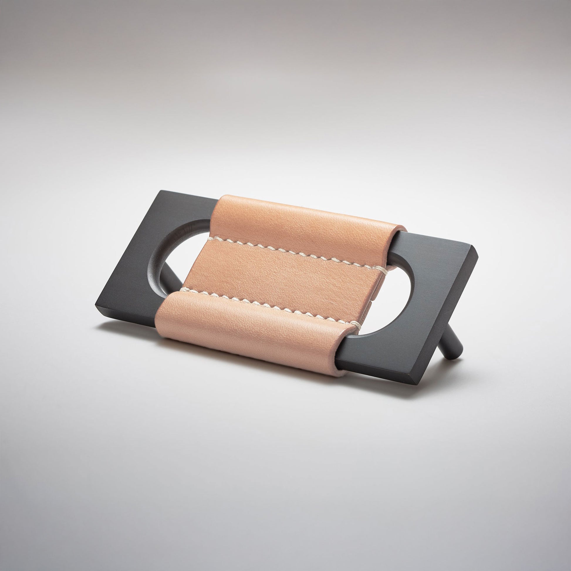 A modern rectangular metal cabinet pull with a central cutout, featuring a light tan leather strap stitched across the top, shown on a clean grey studio background.
