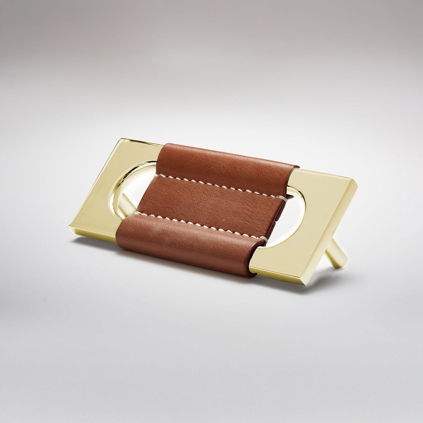 A modern rectangular metal cabinet pull with a central cutout, featuring a light brown leather strap stitched across the top, shown on a clean grey studio background.
