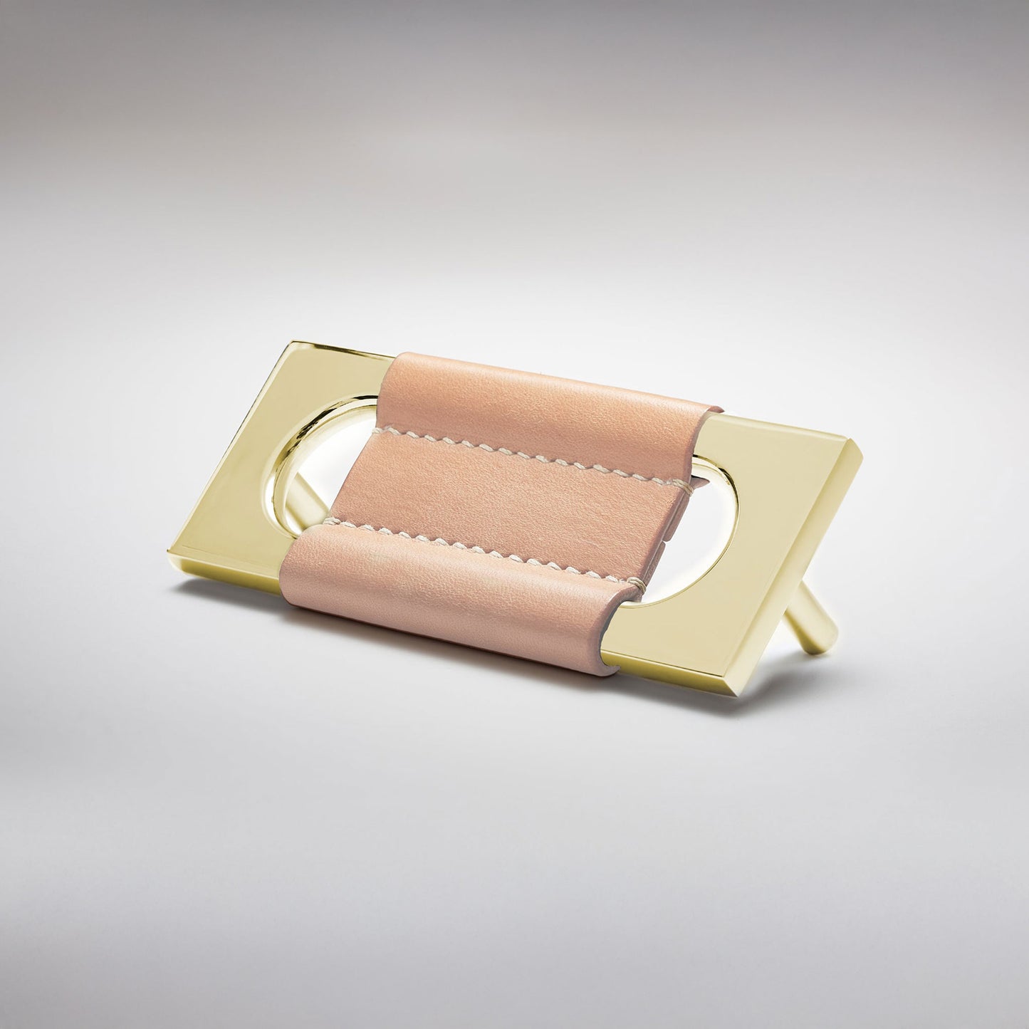 A modern rectangular metal cabinet pull with a central cutout, featuring a light tan leather strap stitched across the top, shown on a clean grey studio background.
