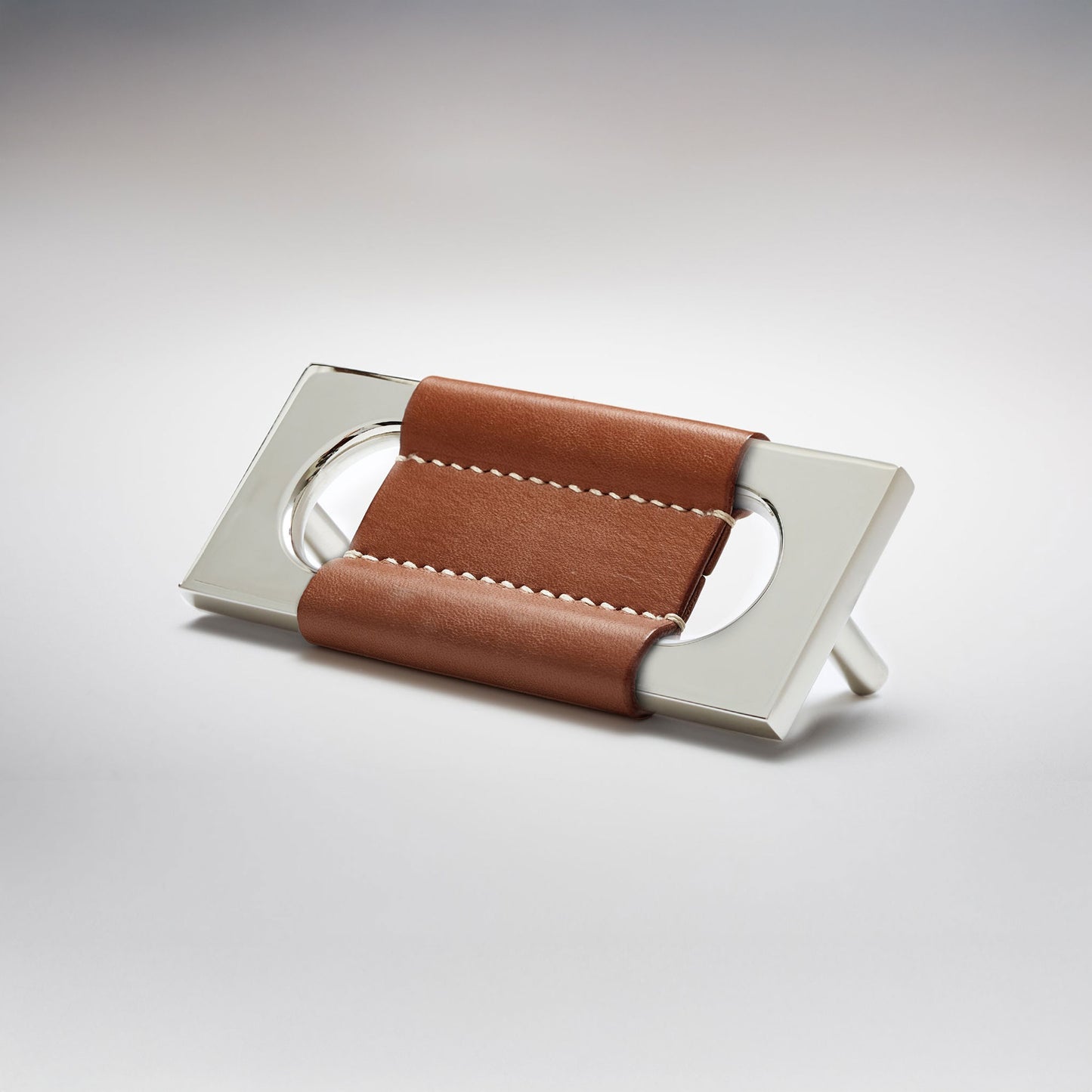 A modern rectangular metal cabinet pull with a central cutout, featuring a light brown leather strap stitched across the top, shown on a clean grey studio background.