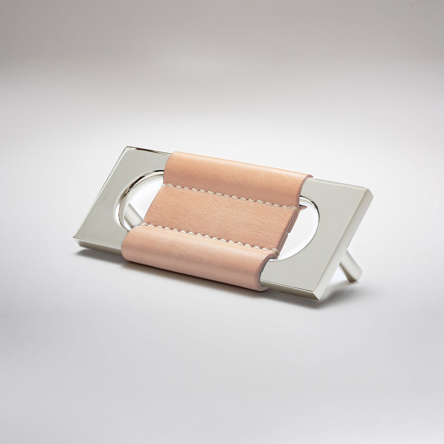 A modern rectangular metal cabinet pull with a central cutout, featuring a light tan leather strap stitched across the top, shown on a clean grey studio background.