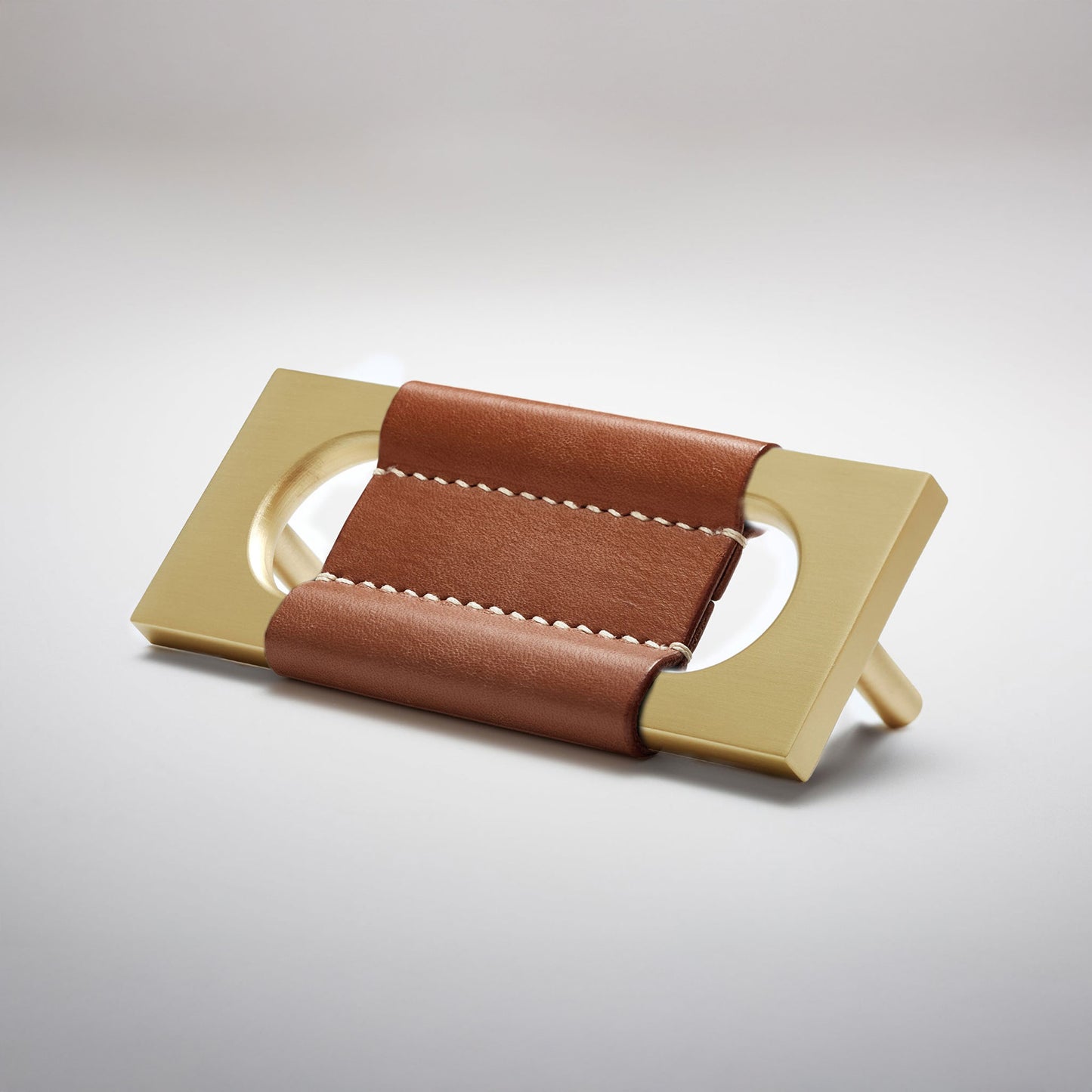 A modern rectangular metal cabinet pull with a central cutout, featuring a light brown leather strap stitched across the top, shown on a clean grey studio background.