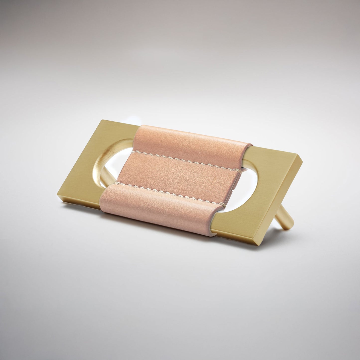 A modern rectangular metal cabinet pull with a central cutout, featuring a light tan leather strap stitched across the top, shown on a clean grey studio background.