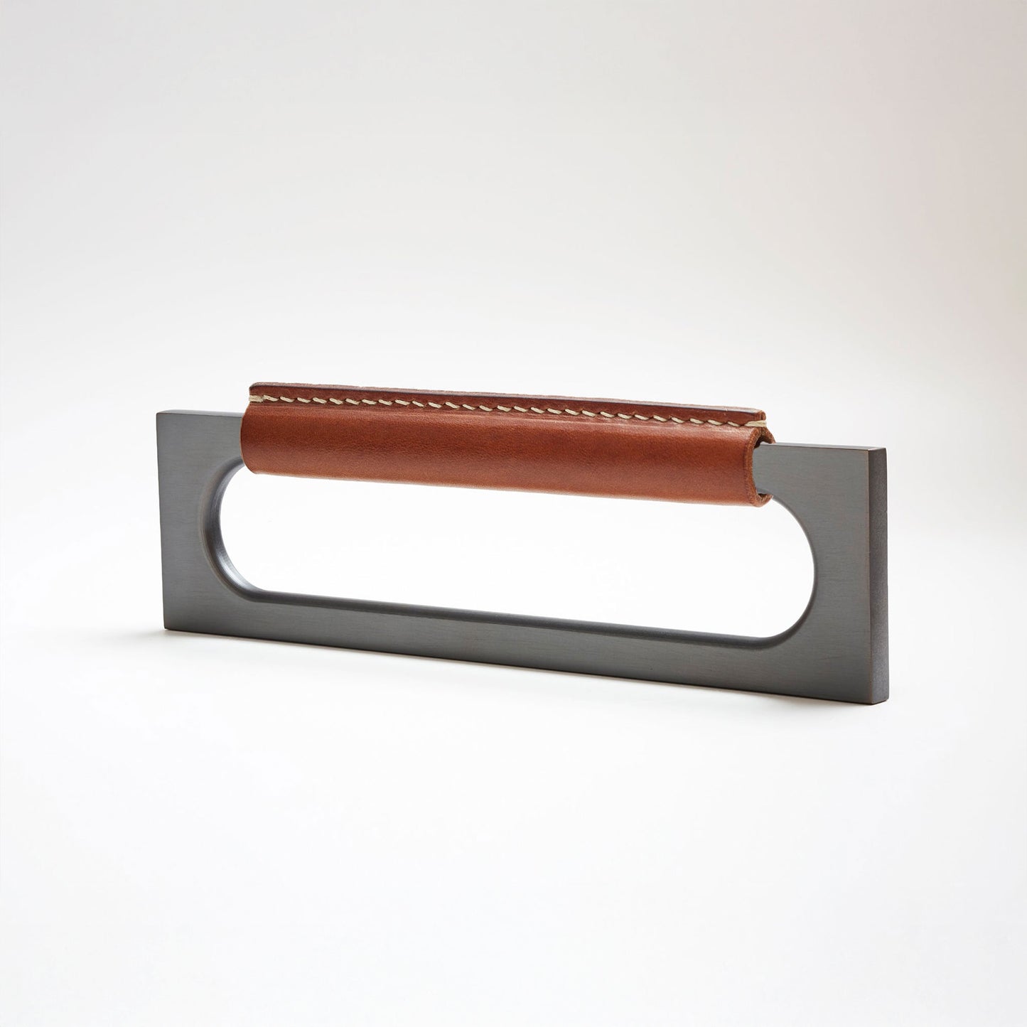 A modern rectangular metal cabinet pull with a central cutout, featuring a light brown leather strap stitched across the top, shown on a clean grey studio background.