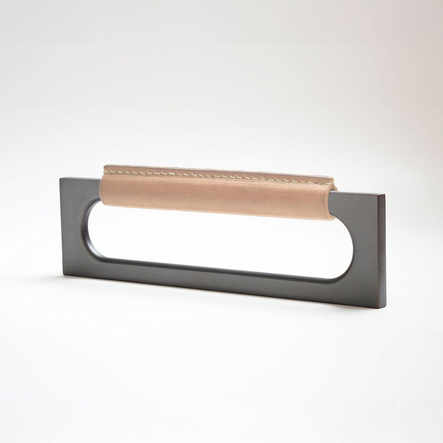 A modern rectangular metal cabinet pull with a central cutout, featuring a light tan leather strap stitched across the top, shown on a clean grey studio background.