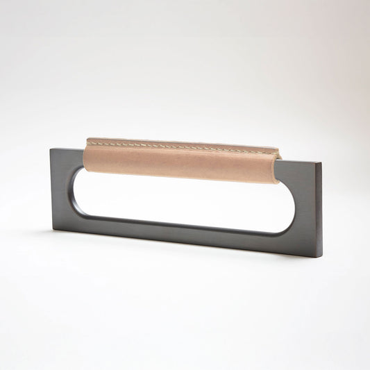 A modern rectangular metal cabinet pull with a central cutout, featuring a light tan leather strap stitched across the top, shown on a clean grey studio background.