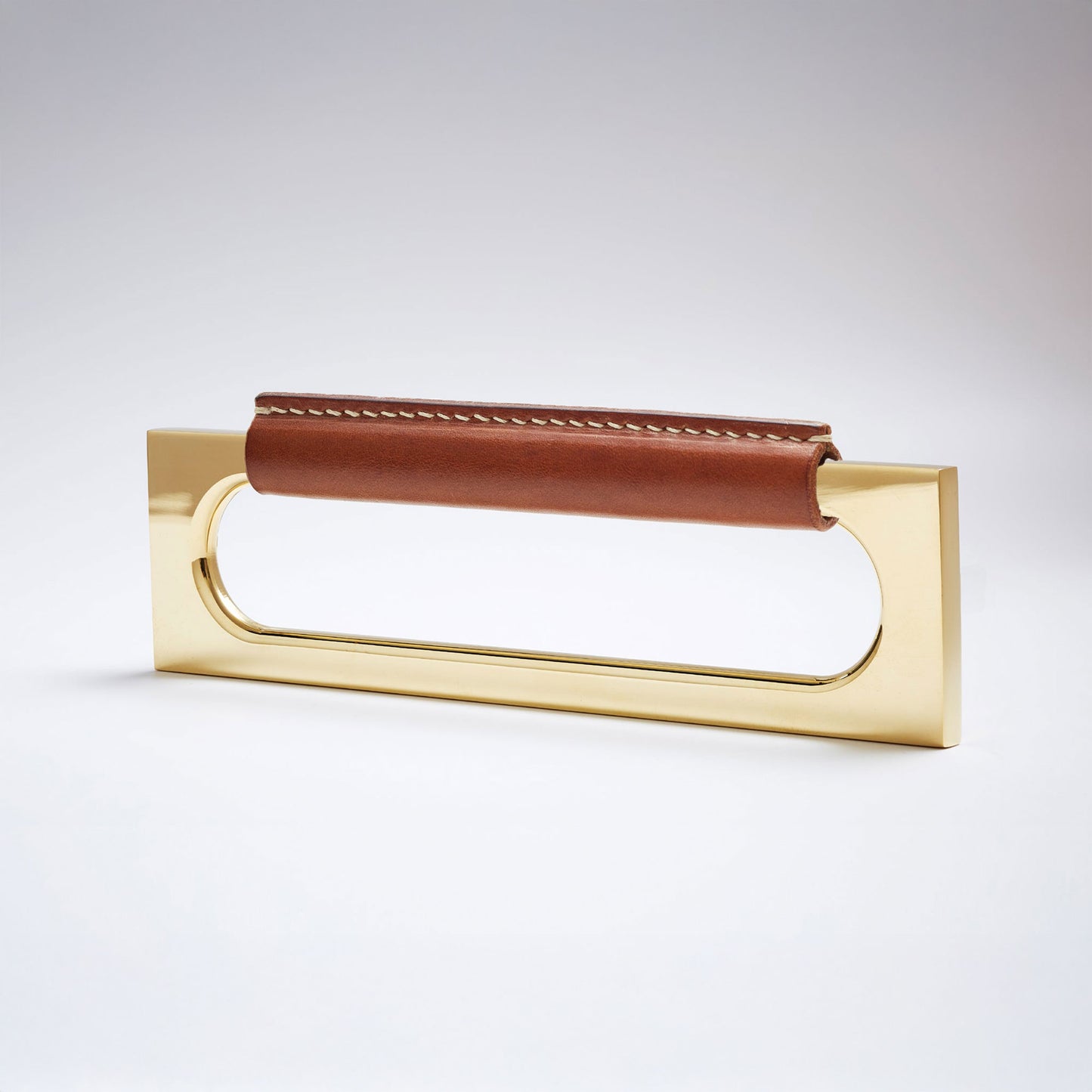 A modern rectangular metal cabinet pull with a central cutout, featuring a light brown leather strap stitched across the top, shown on a clean grey studio background.