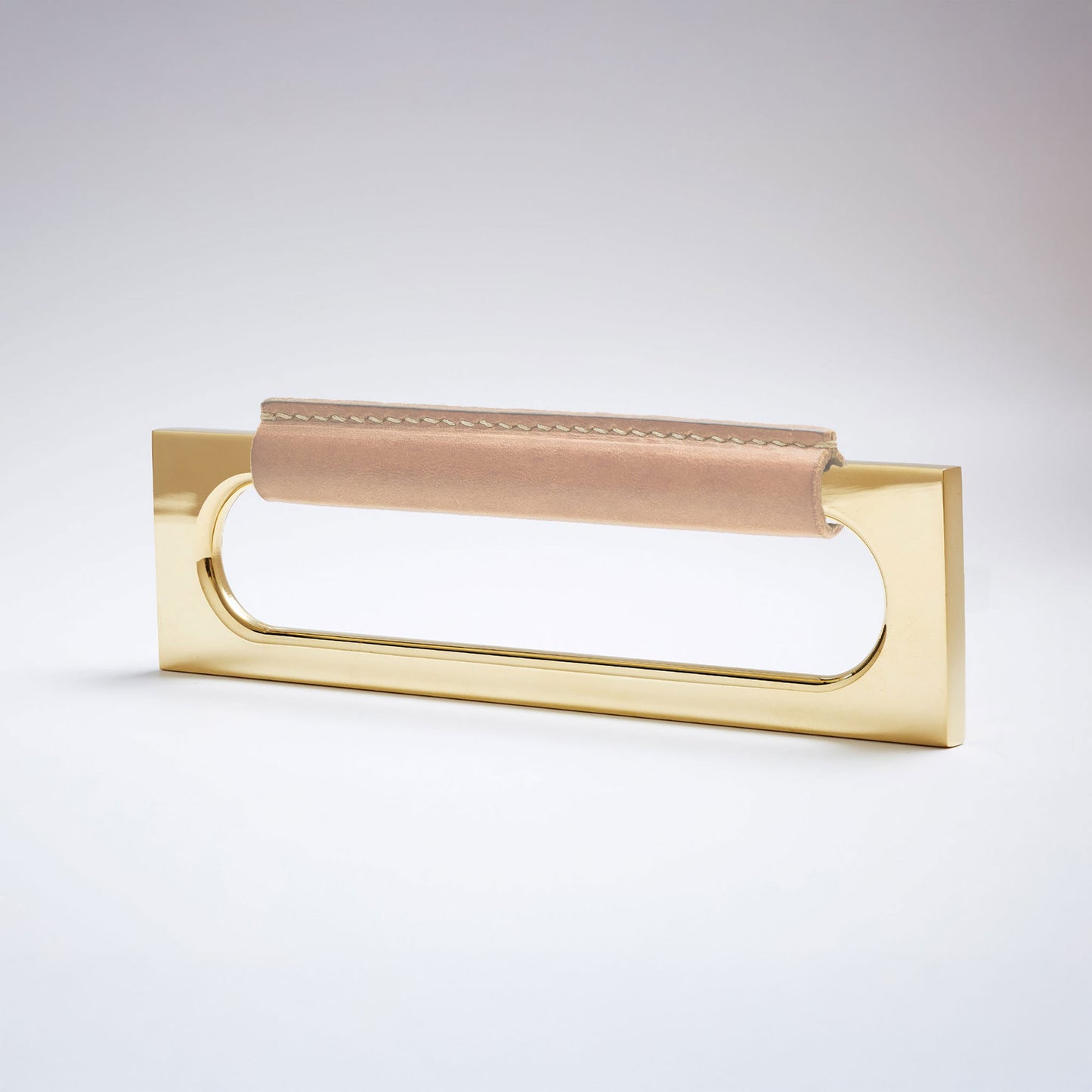 A modern rectangular metal cabinet pull with a central cutout, featuring a light tan leather strap stitched across the top, shown on a clean grey studio background.