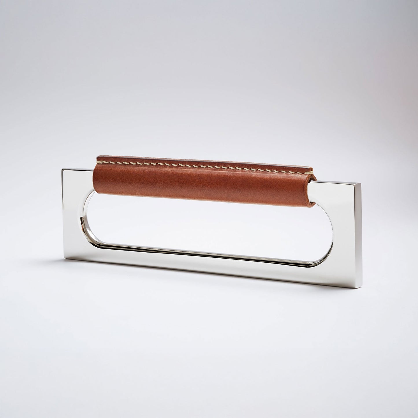 A modern rectangular metal cabinet pull with a central cutout, featuring a light brown leather strap stitched across the top, shown on a clean grey studio background.