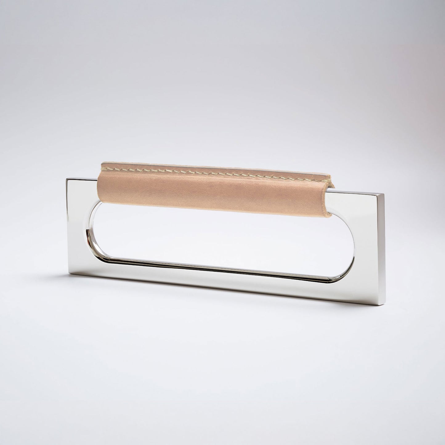 A modern rectangular metal cabinet pull with a central cutout, featuring a light tan leather strap stitched across the top, shown on a clean grey studio background.