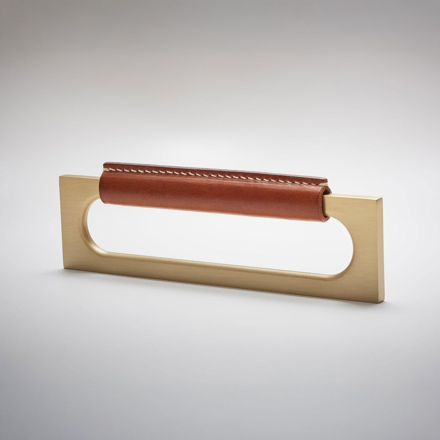 A modern rectangular metal cabinet pull with a central cutout, featuring a light brown leather strap stitched across the top, shown on a clean grey studio background.