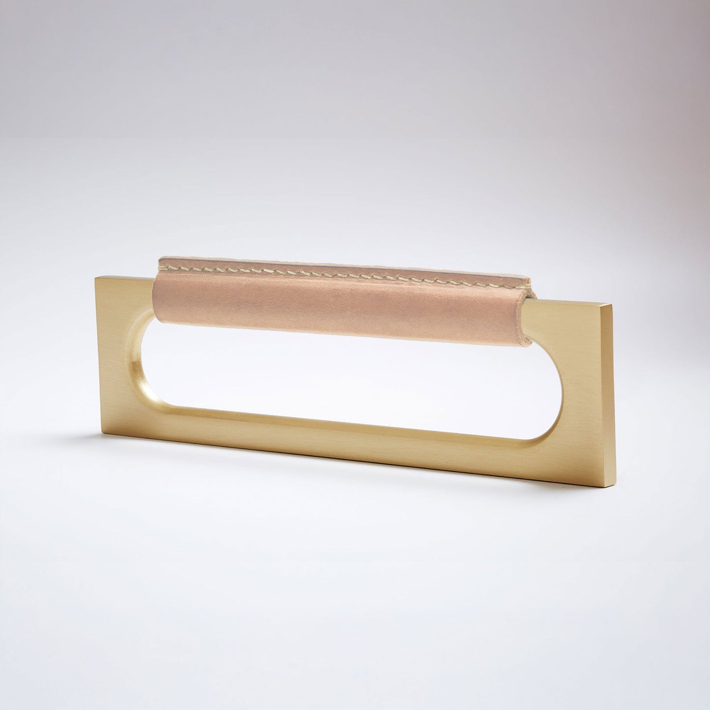A modern rectangular metal cabinet pull with a central cutout, featuring a light tan leather strap stitched across the top, shown on a clean grey studio background.