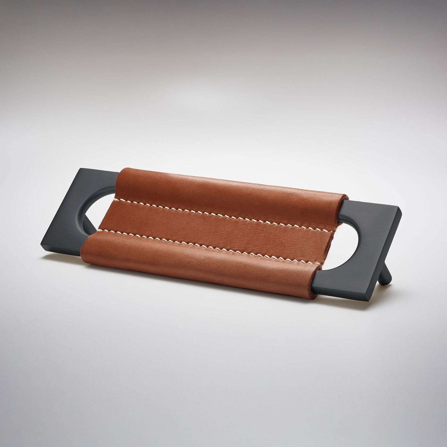 A modern rectangular metal cabinet pull with a central cutout, featuring a light brown leather strap stitched across the top, shown on a clean grey studio background.