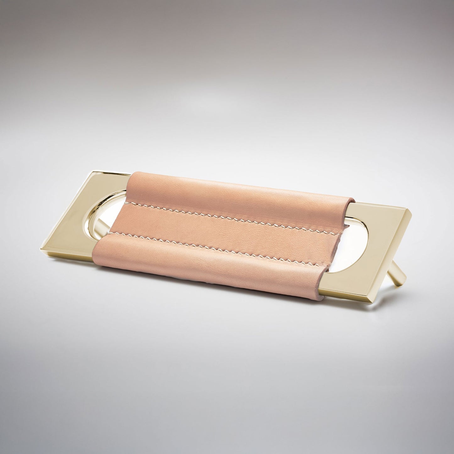 A modern rectangular metal cabinet pull with a central cutout, featuring a light tan leather strap stitched across the top, shown on a clean grey studio background.