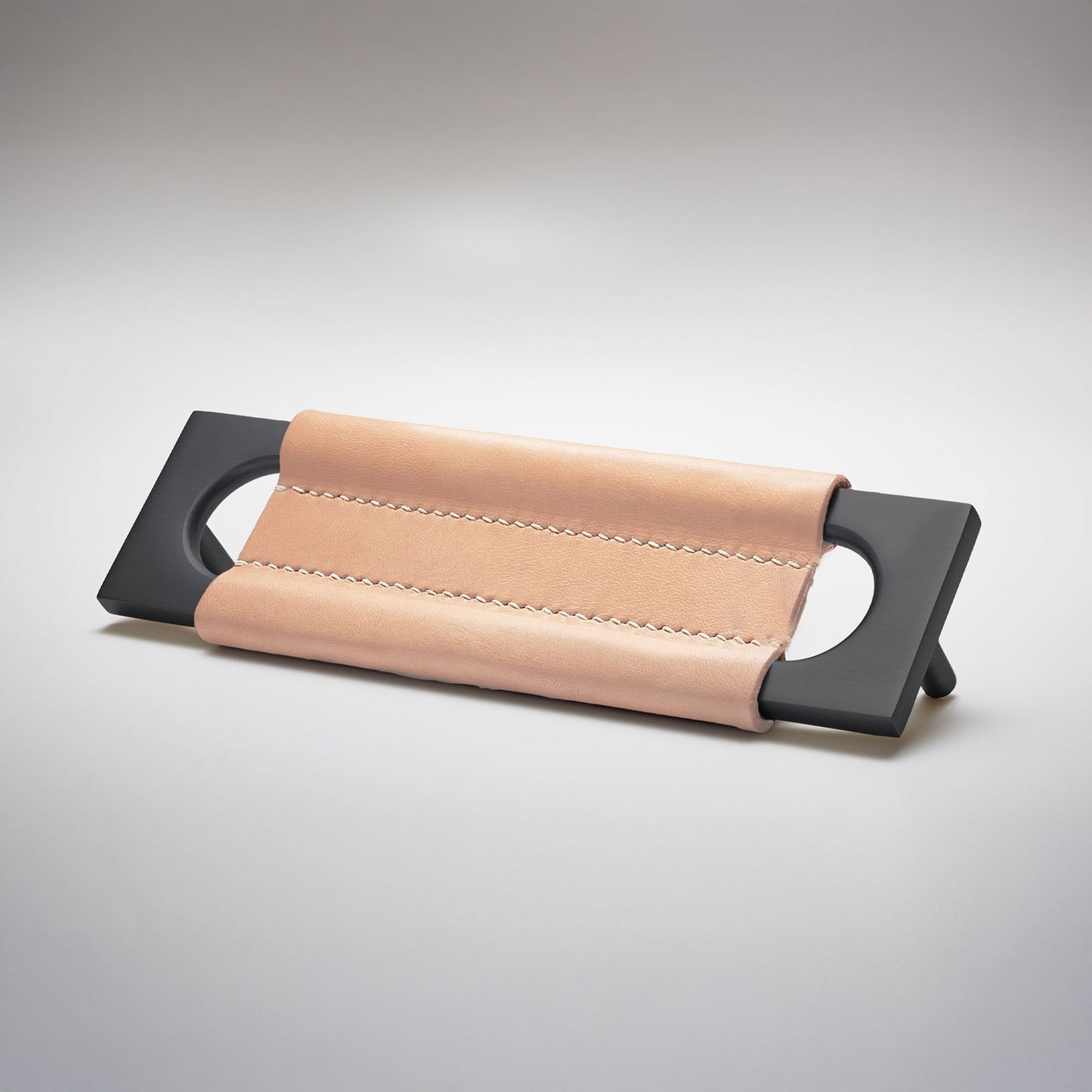 A modern rectangular metal cabinet pull with a central cutout, featuring a light tan leather strap stitched across the top, shown on a clean grey studio background.