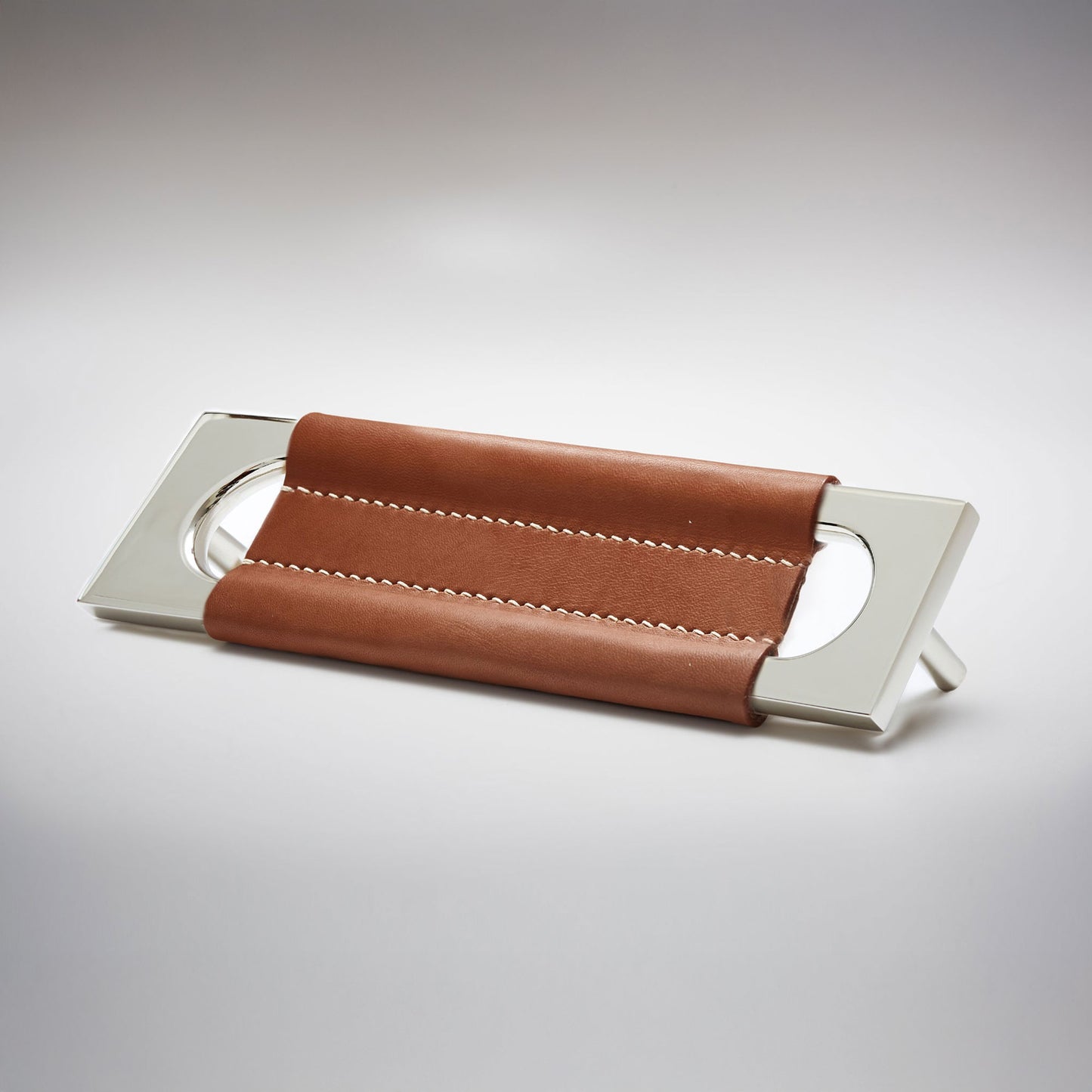 A modern rectangular metal cabinet pull with a central cutout, featuring a light brown leather strap stitched across the top, shown on a clean grey studio background.
