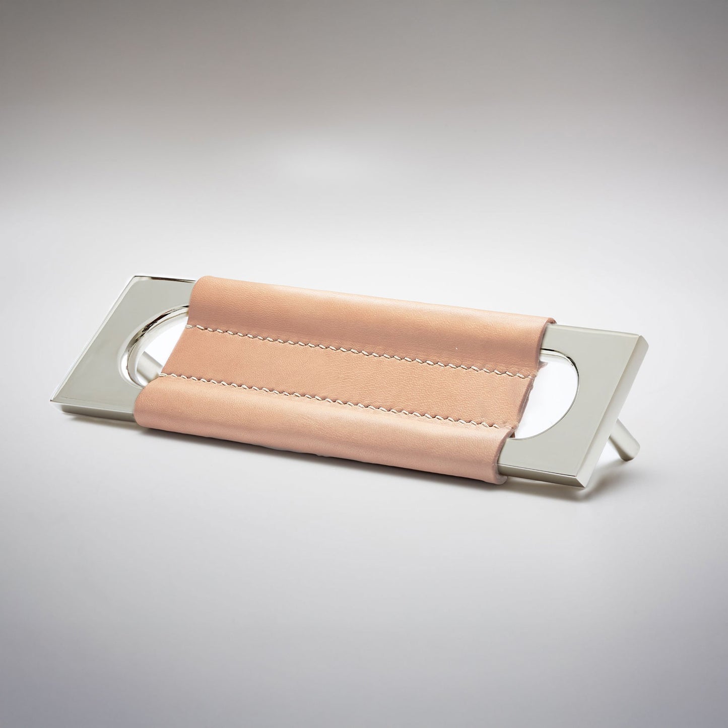 A modern rectangular metal cabinet pull with a central cutout, featuring a light tan leather strap stitched across the top, shown on a clean grey studio background.