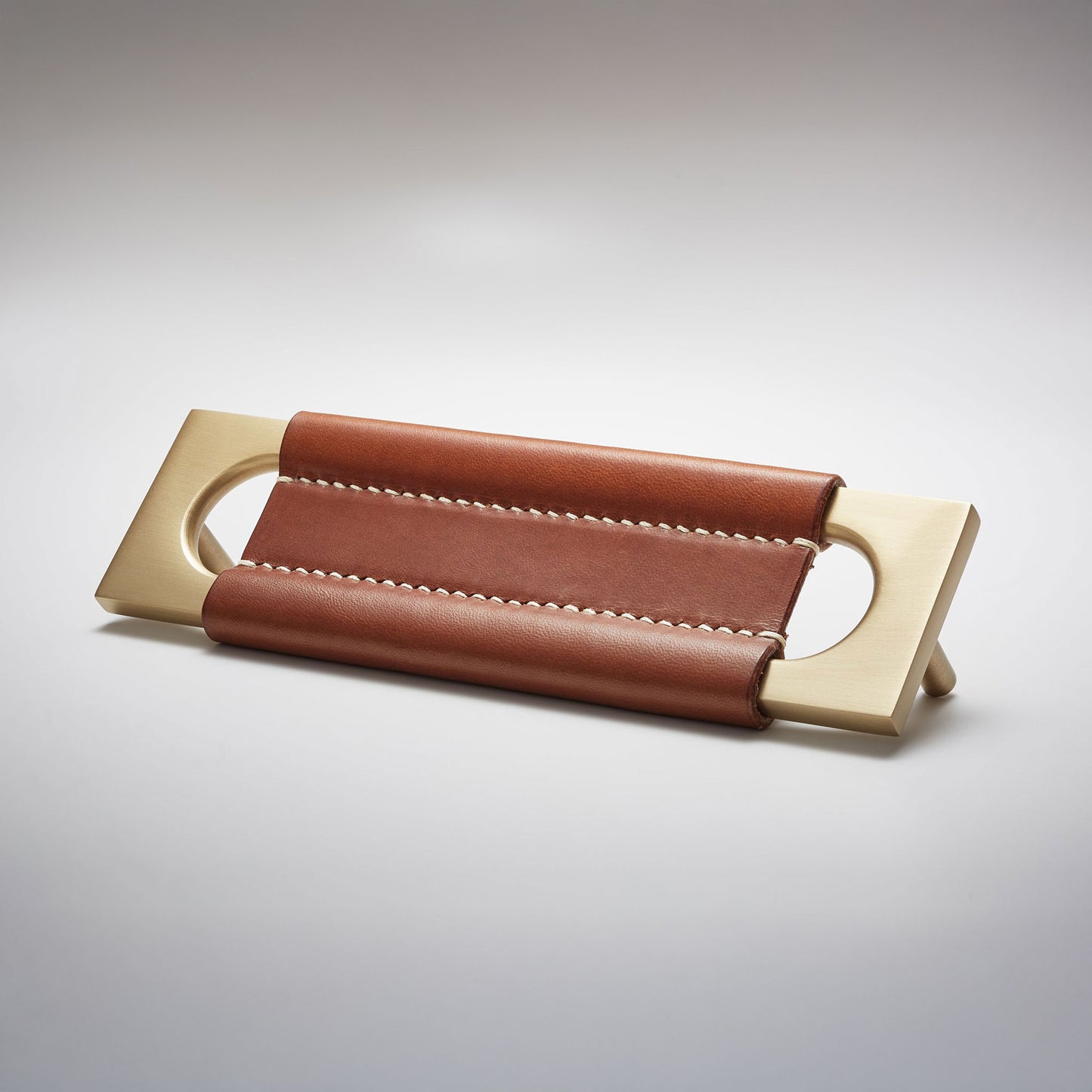 A modern rectangular metal cabinet pull with a central cutout, featuring a light brown leather strap stitched across the top, shown on a clean grey studio background.