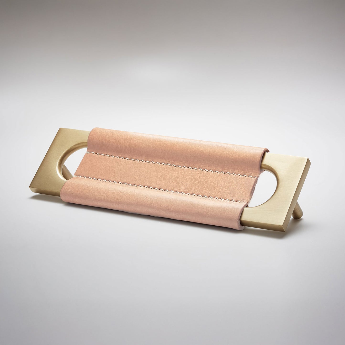 A modern rectangular metal cabinet pull with a central cutout, featuring a light tan leather strap stitched across the top, shown on a clean grey studio background.