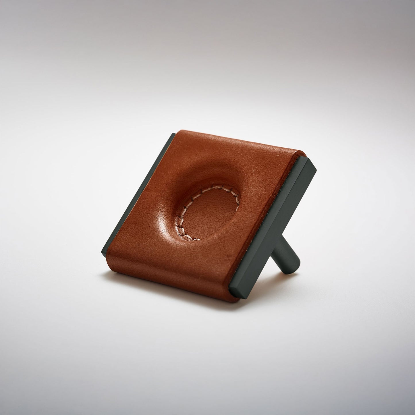 A dark leather and metal square cabinet knob on a clean greay background