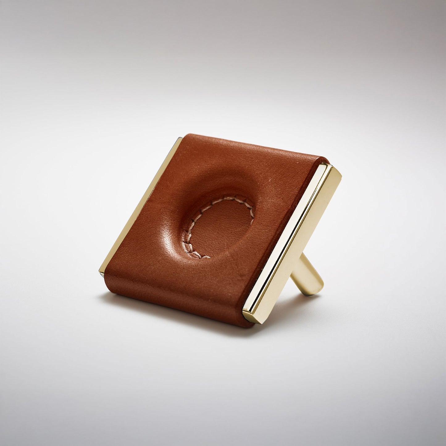 A dark leather and metal square cabinet knob on a clean greay background