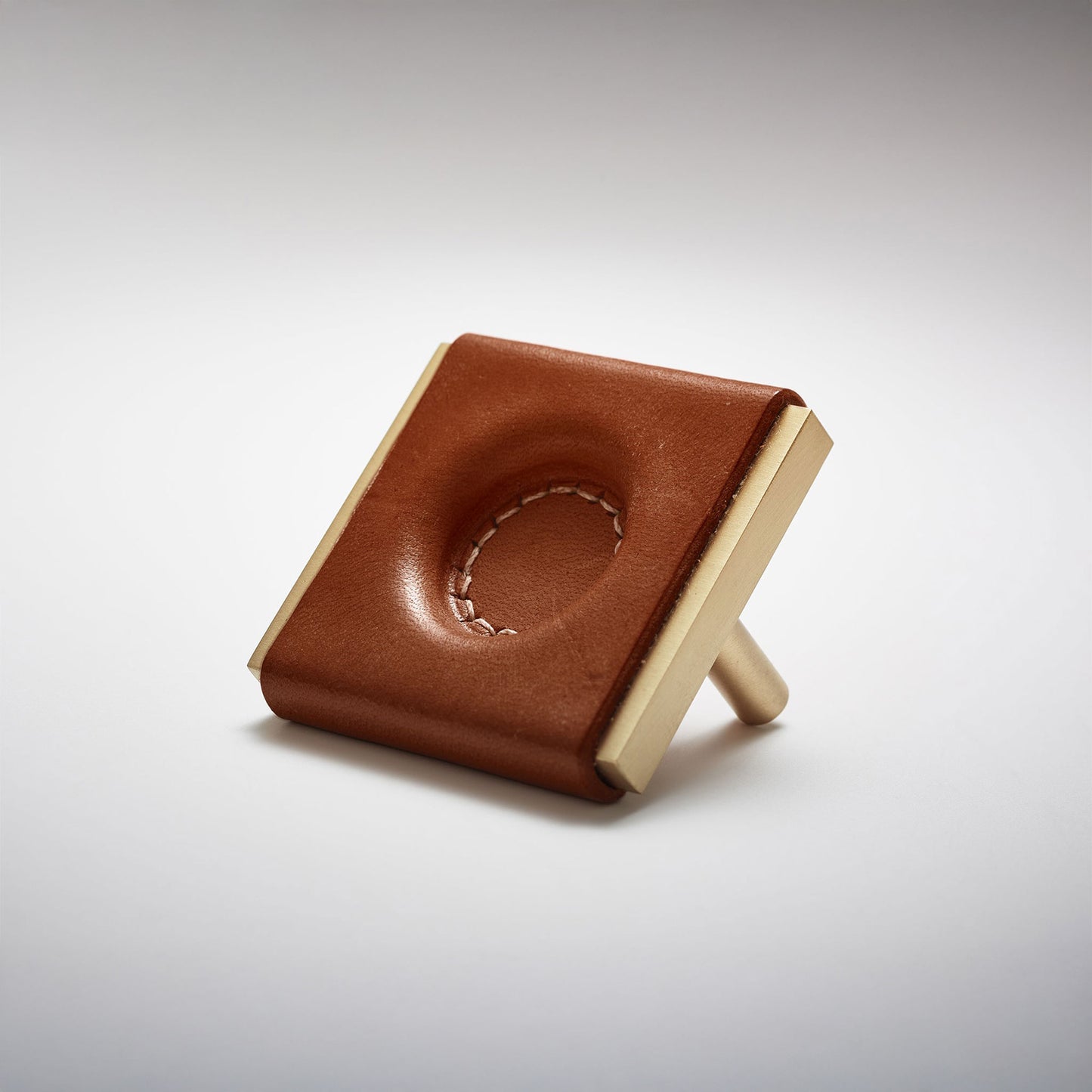A dark leather and metal square cabinet knob on a clean greay background