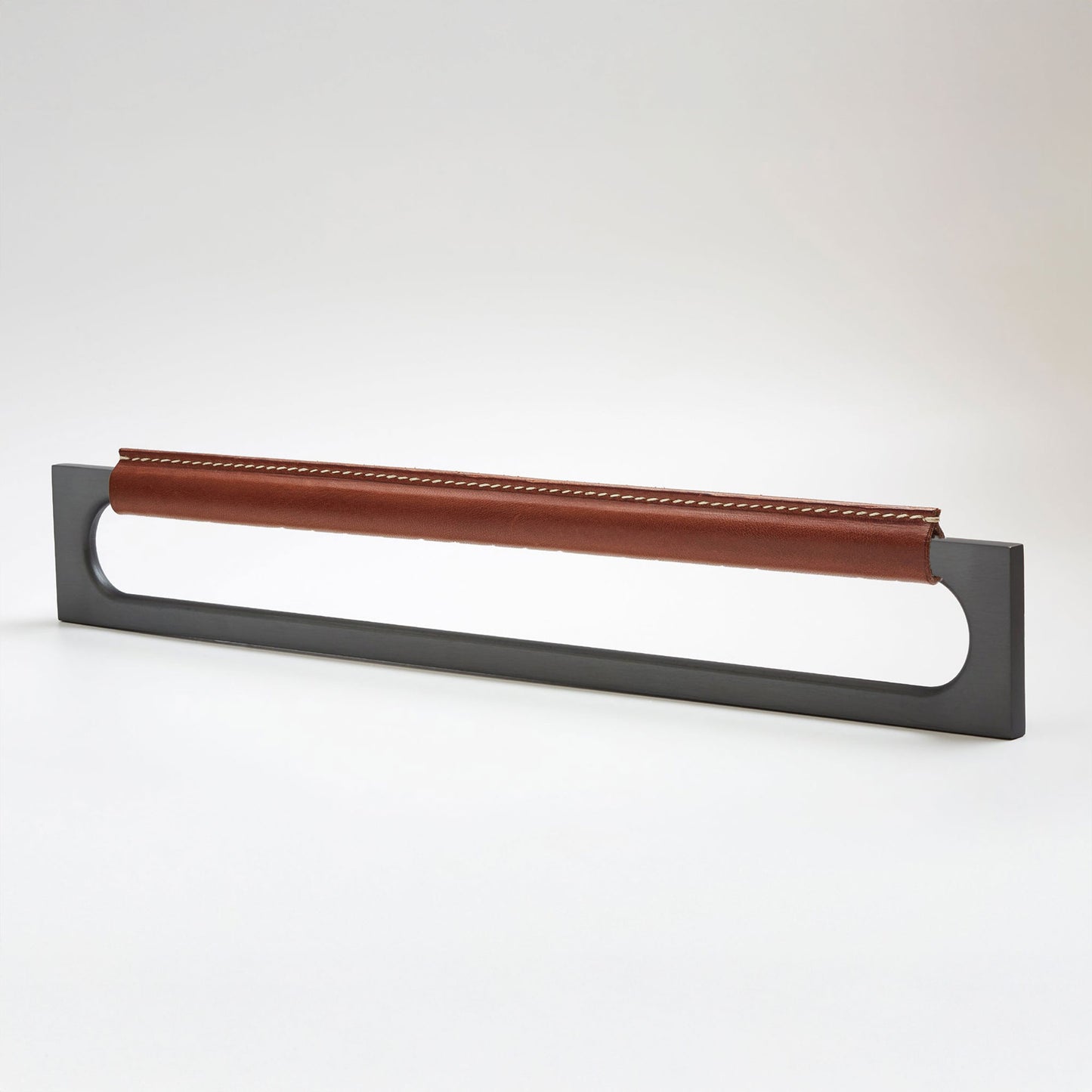 A modern rectangular metal cabinet pull with a central cutout, featuring a light brown leather strap stitched across the top, shown on a clean grey studio background.