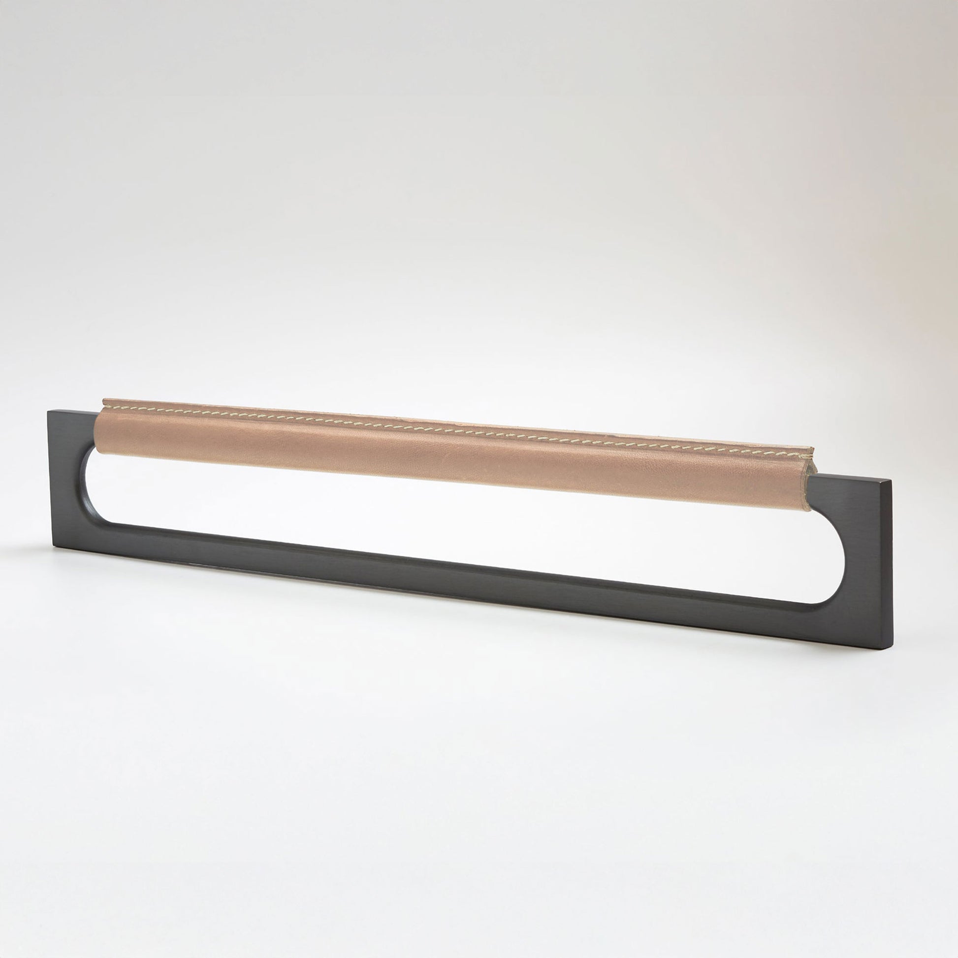A modern rectangular metal cabinet pull with a central cutout, featuring a light tan leather strap stitched across the top, shown on a clean grey studio background.