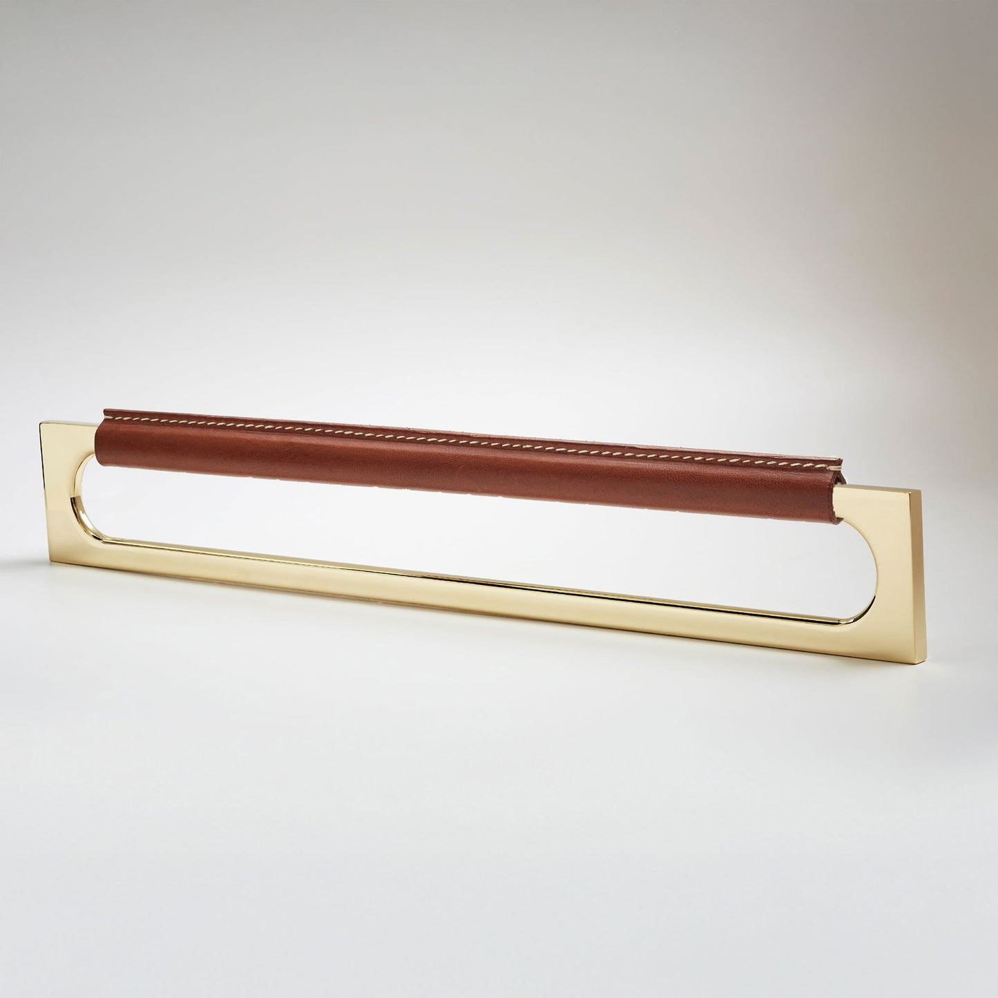 A modern rectangular metal cabinet pull with a central cutout, featuring a light brown leather strap stitched across the top, shown on a clean grey studio background.