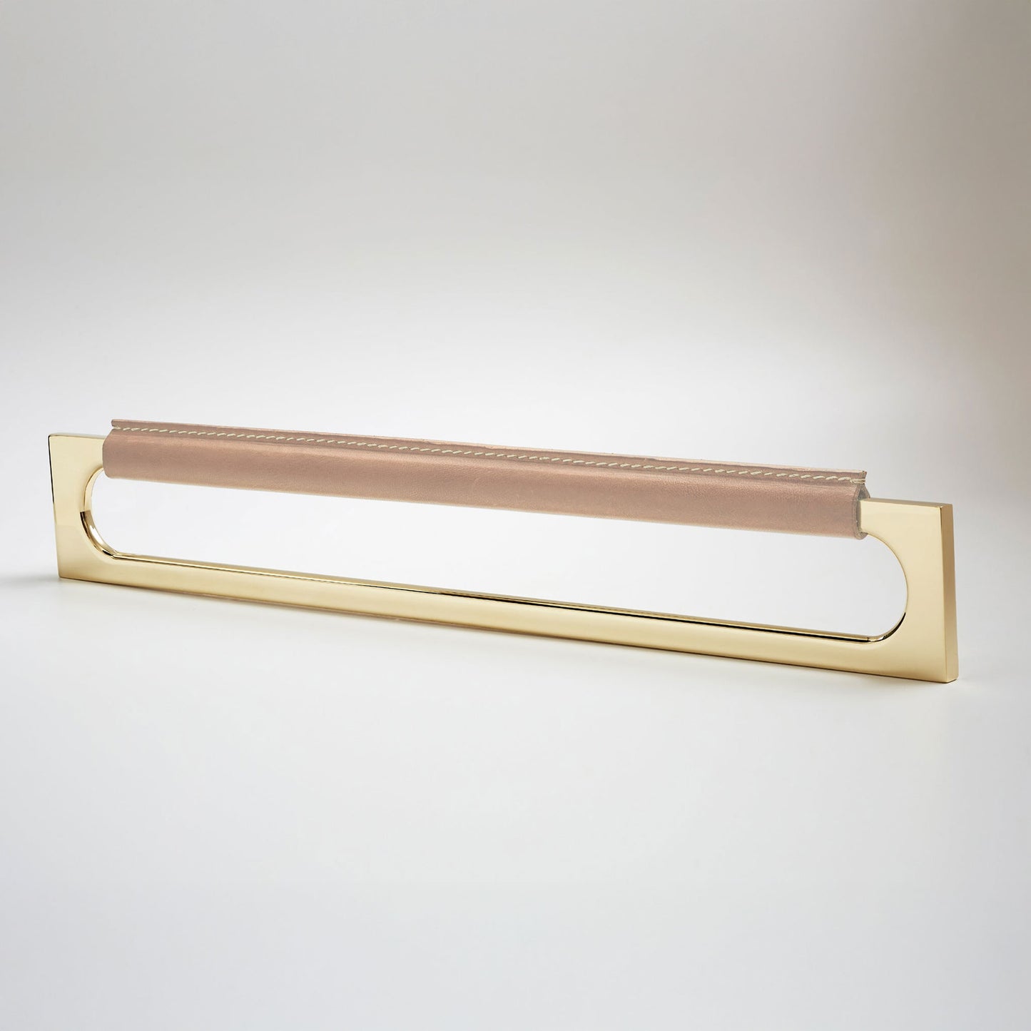 A modern rectangular metal cabinet pull with a central cutout, featuring a light tan leather strap stitched across the top, shown on a clean grey studio background.