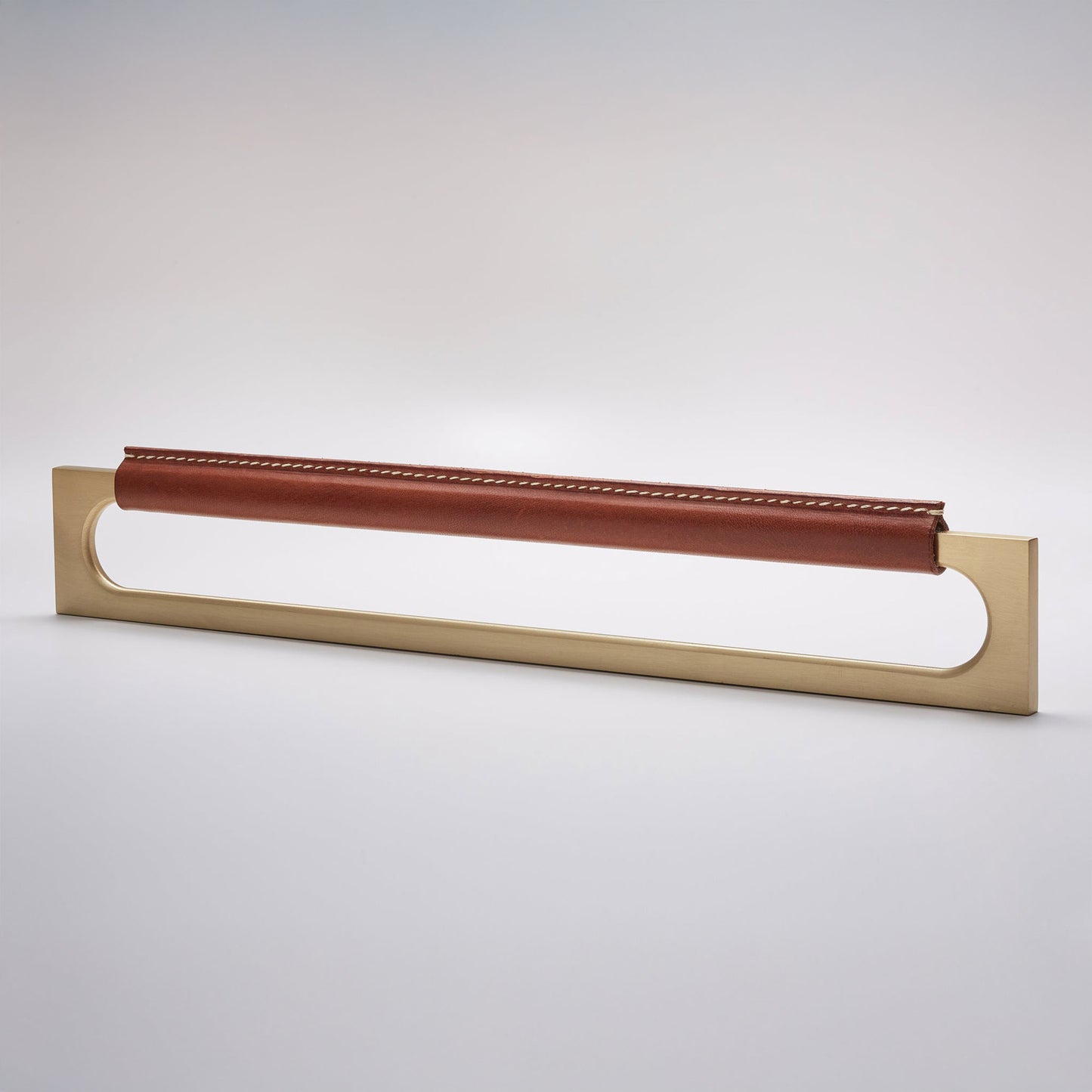 A modern rectangular metal cabinet pull with a central cutout, featuring a light brown leather strap stitched across the top, shown on a clean grey studio background.