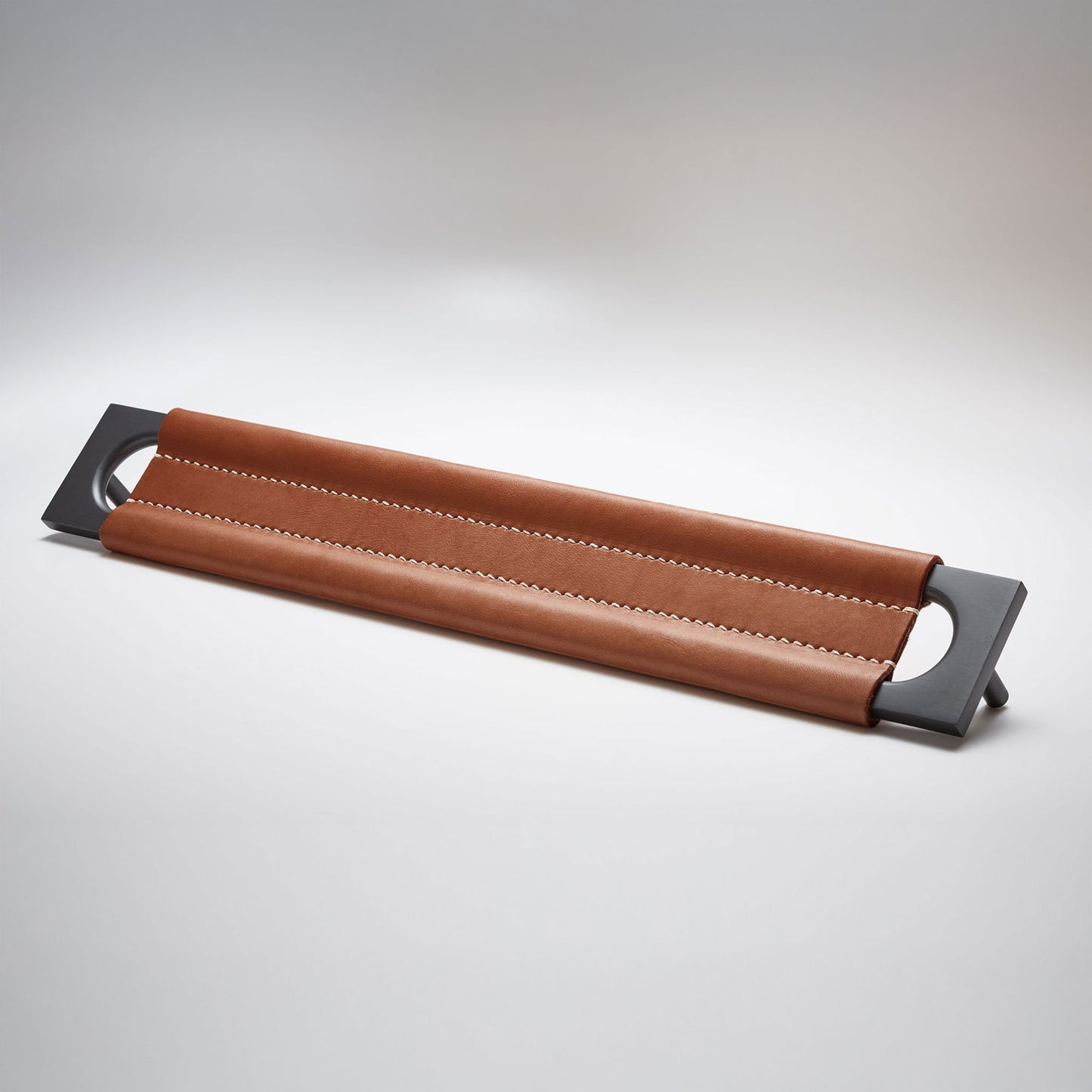 A modern rectangular metal cabinet pull with a central cutout, featuring a light brown leather strap stitched across the top, shown on a clean grey studio background.