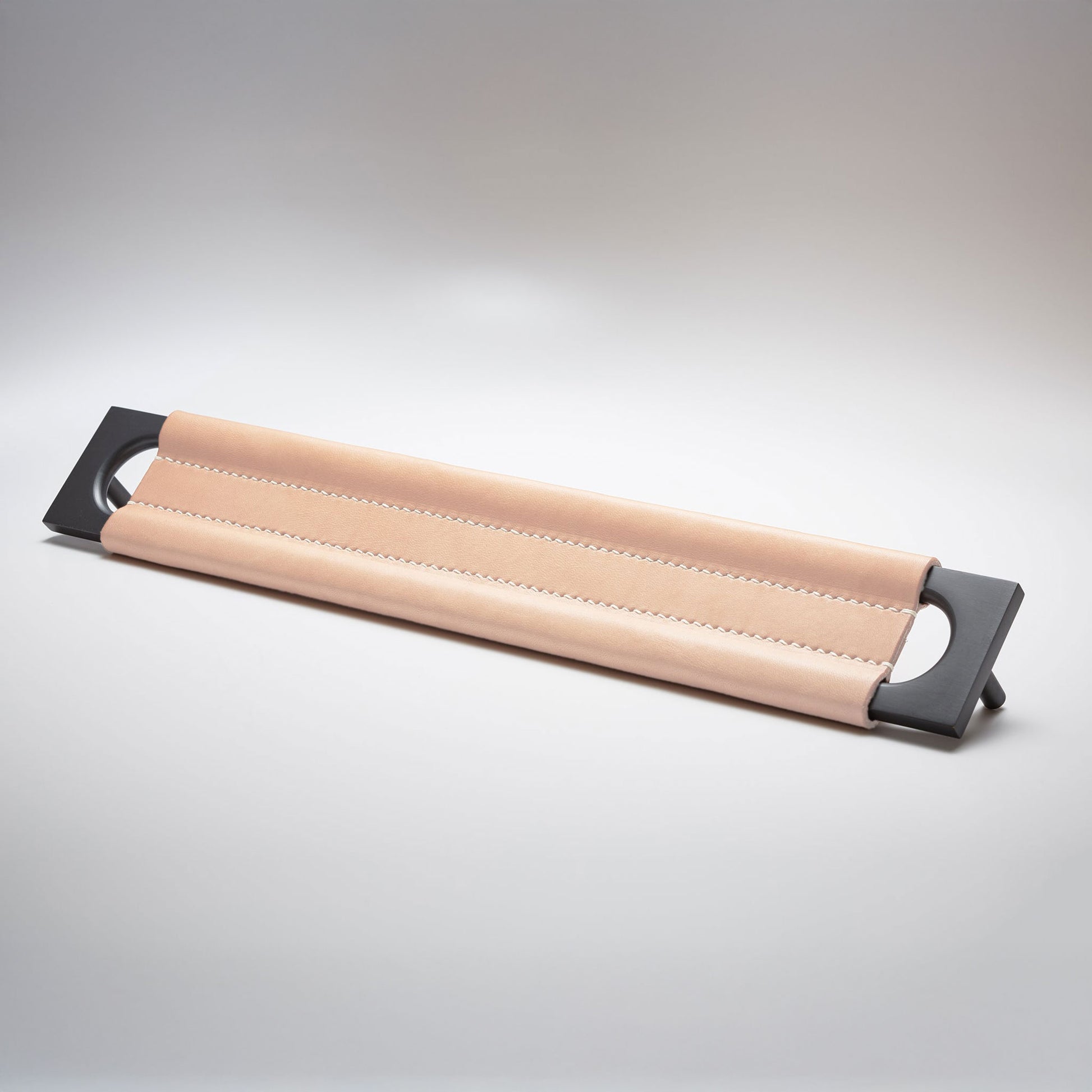 A modern rectangular metal cabinet pull with a central cutout, featuring a light tan leather strap stitched across the top, shown on a clean grey studio background.