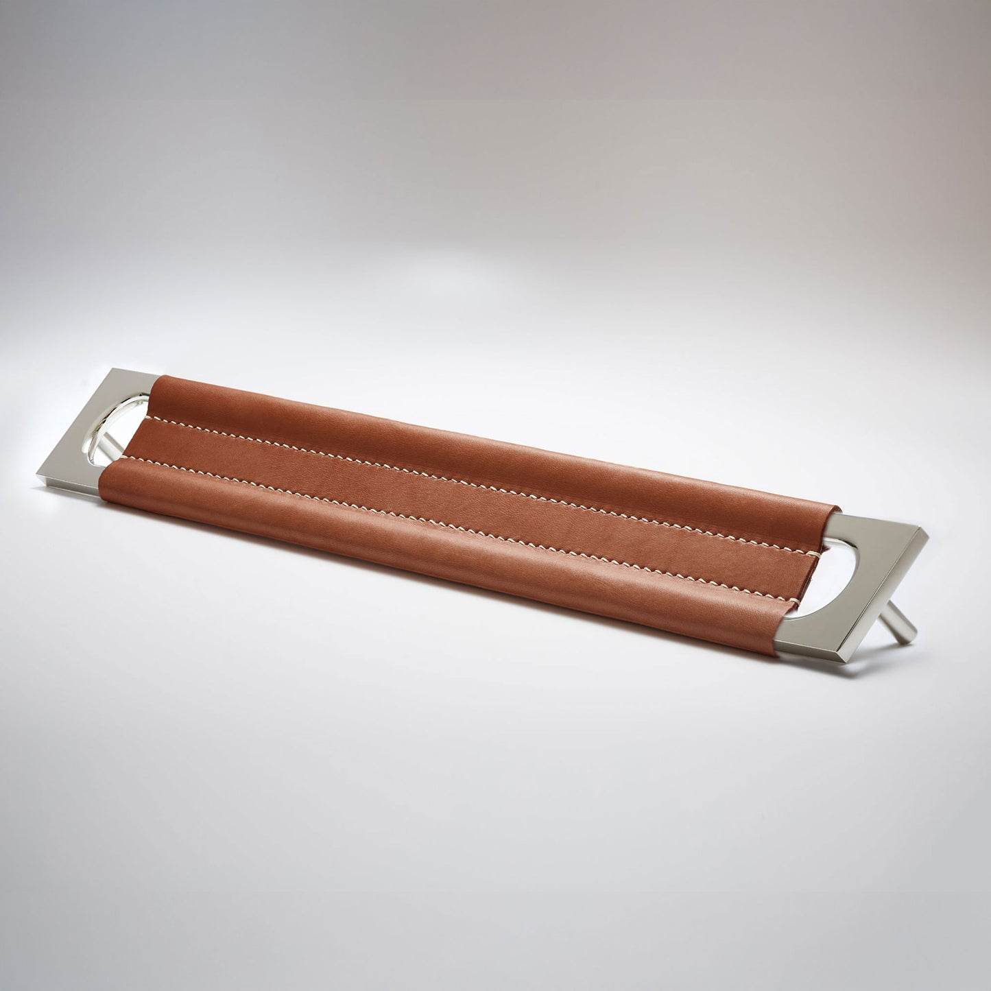 A modern rectangular metal cabinet pull with a central cutout, featuring a light brown leather strap stitched across the top, shown on a clean grey studio background.