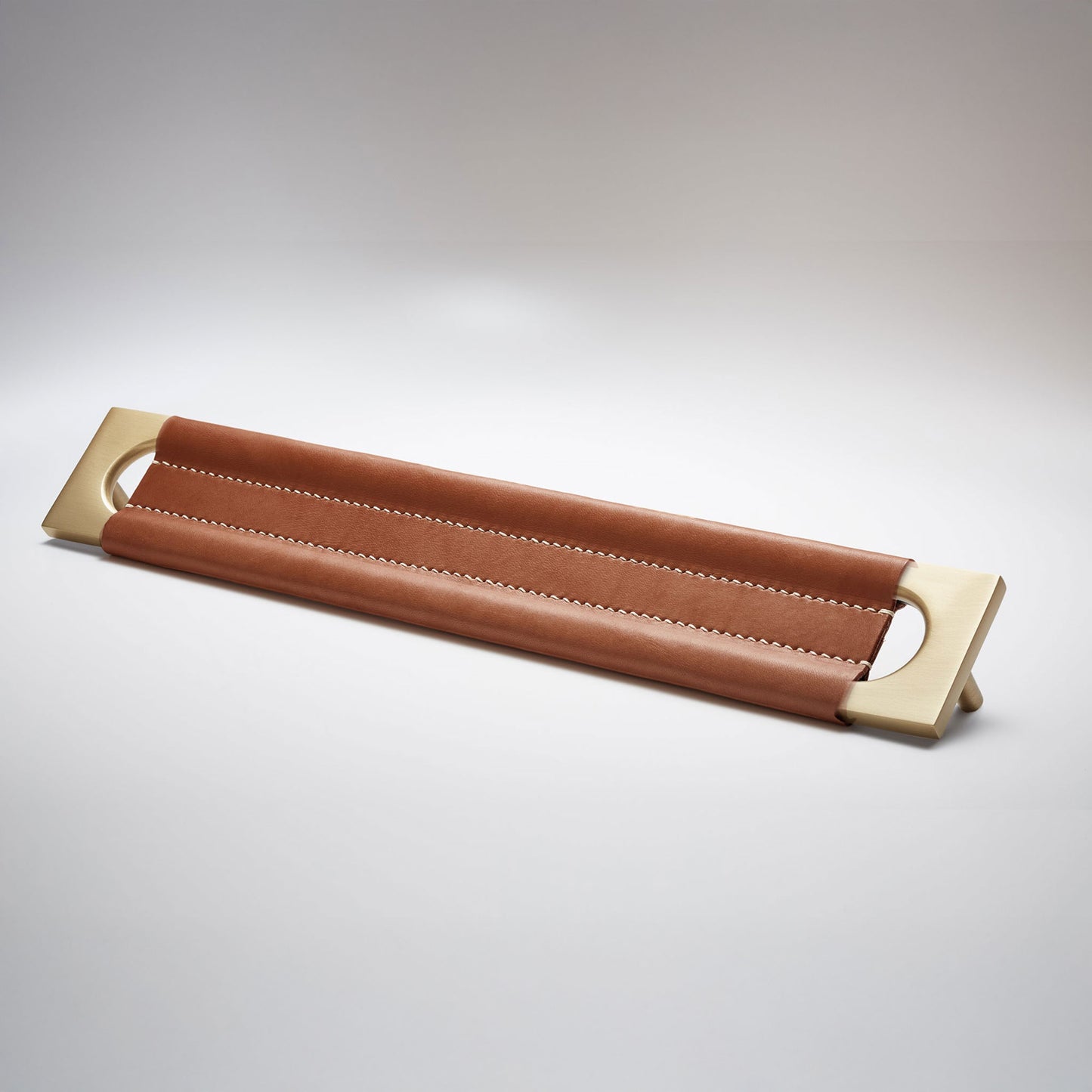 A modern rectangular metal cabinet pull with a central cutout, featuring a light brown leather strap stitched across the top, shown on a clean grey studio background.
