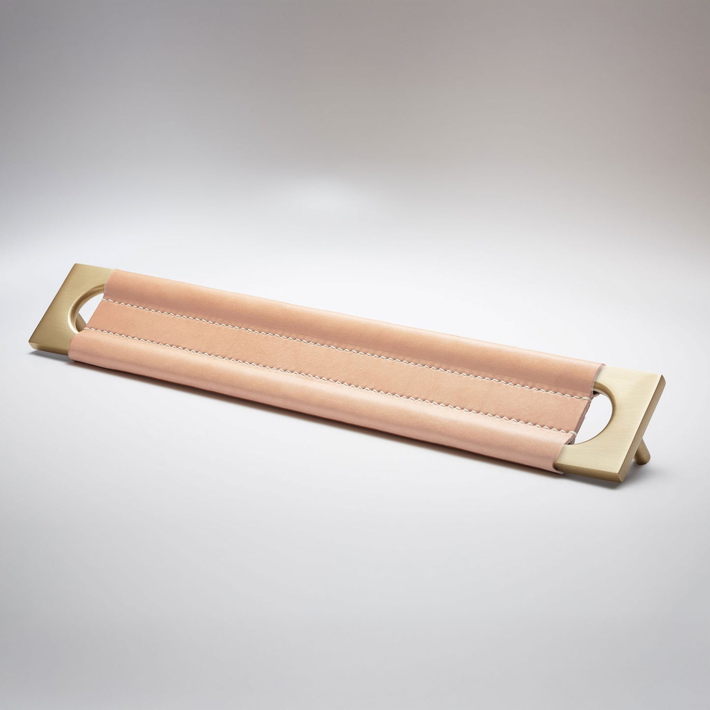 A modern rectangular metal cabinet pull with a central cutout, featuring a light tan leather strap stitched across the top, shown on a clean grey studio background.