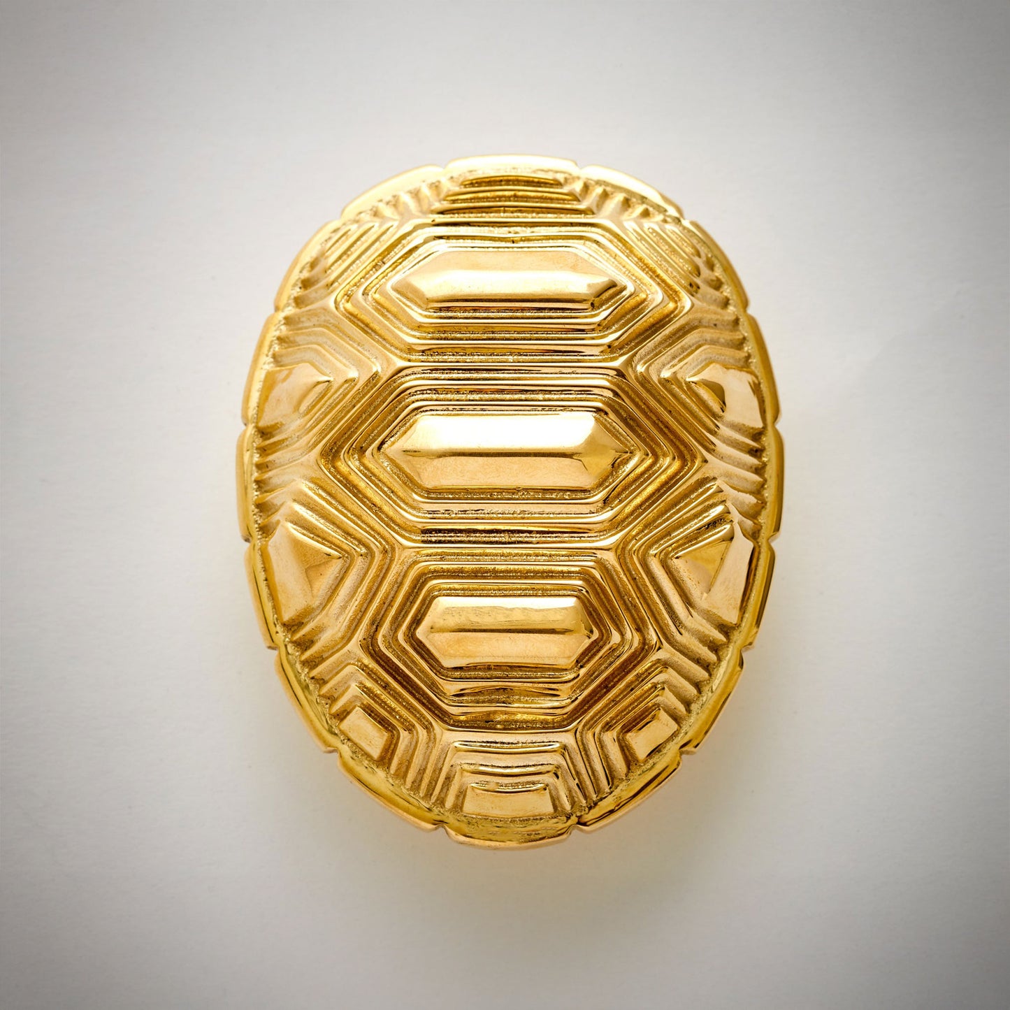 An oval-shaped decorative cabinet pull featuring a textured polished natural brass geometric pattern, on a neutral light gray background.