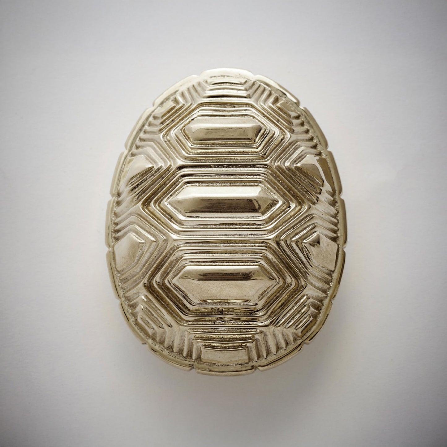An oval-shaped decorative cabinet pull featuring a textured polished nickel geometric pattern, on a neutral light gray background.