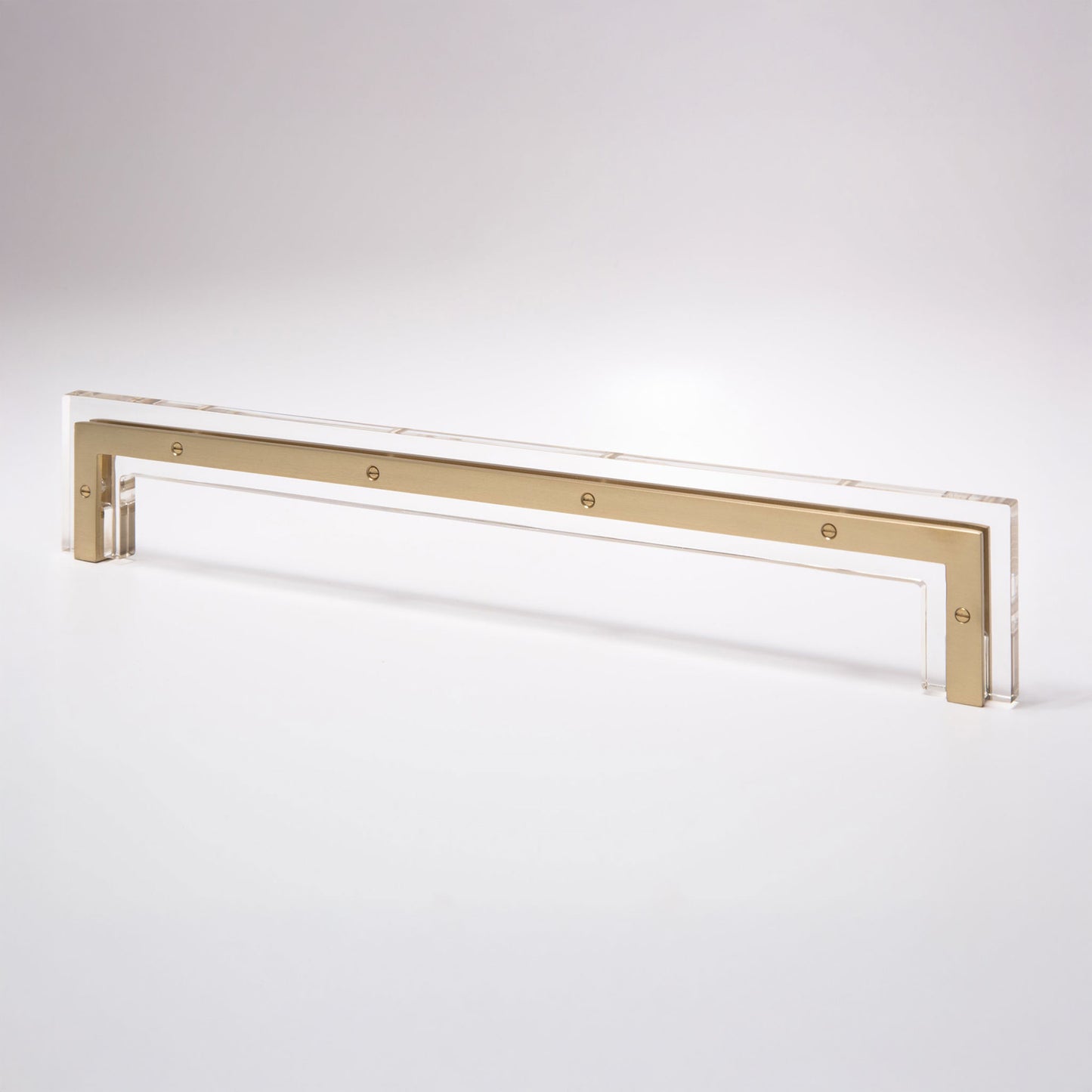 A modern horizontal cabinet pull handle featuring a brushed gold metal bar set within a clear acrylic frame, shown on a seamless light gray background.