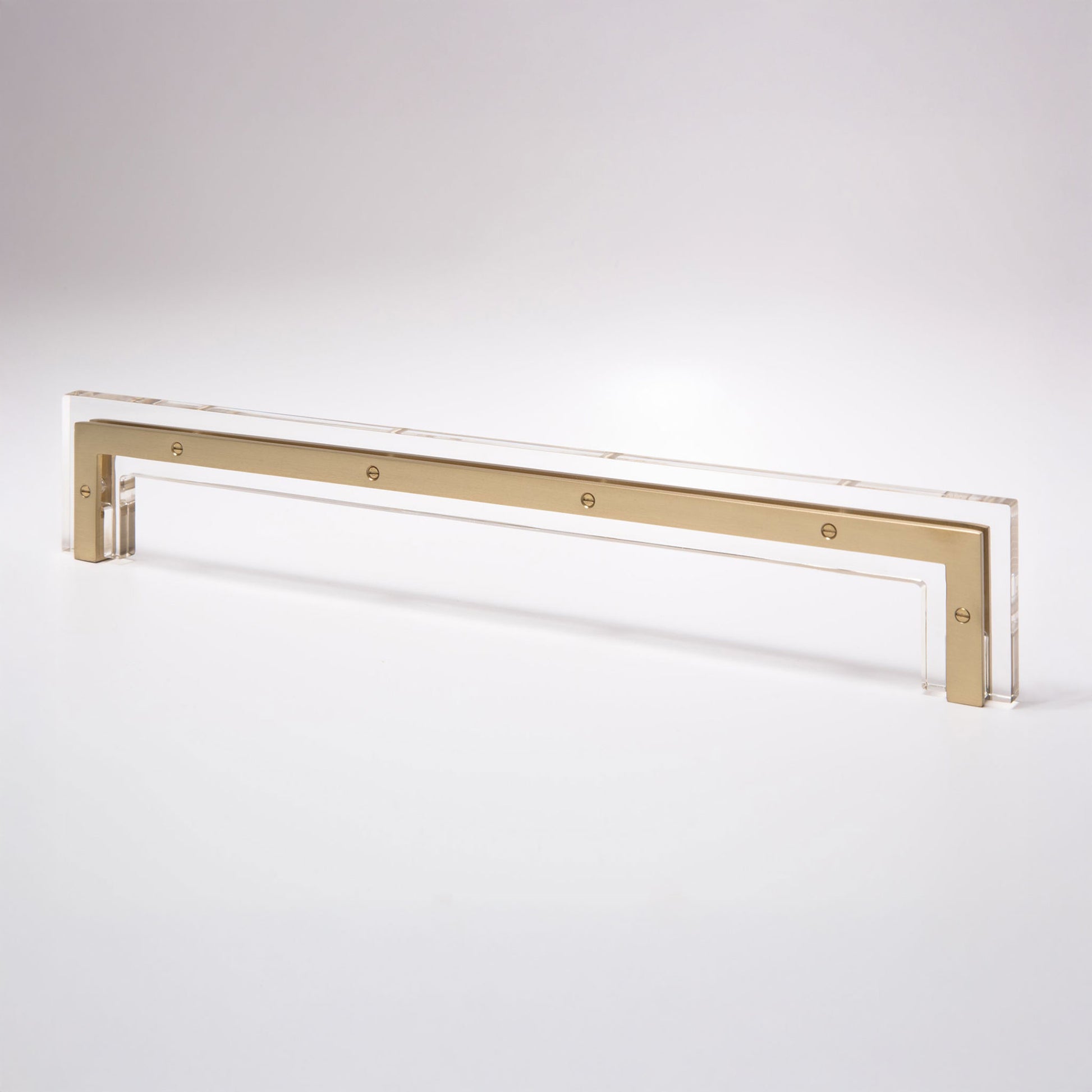 A modern horizontal cabinet pull handle featuring a brushed gold metal bar set within a clear acrylic frame, shown on a seamless light gray background.