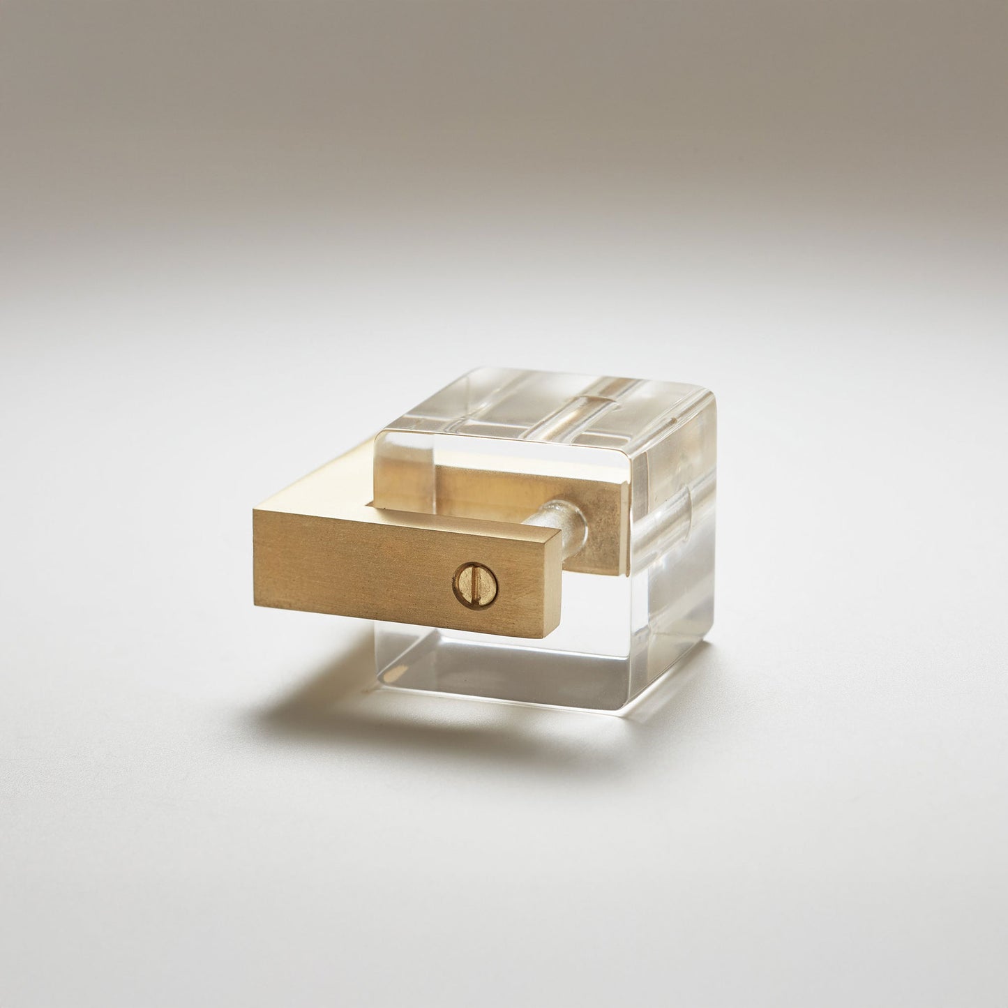 A modern acrylic and brasscabinet knob, featuring a transparent clear acrylic cube with a brushed brass rectangular handle and a small screw detail, set against a neutral light gray background.