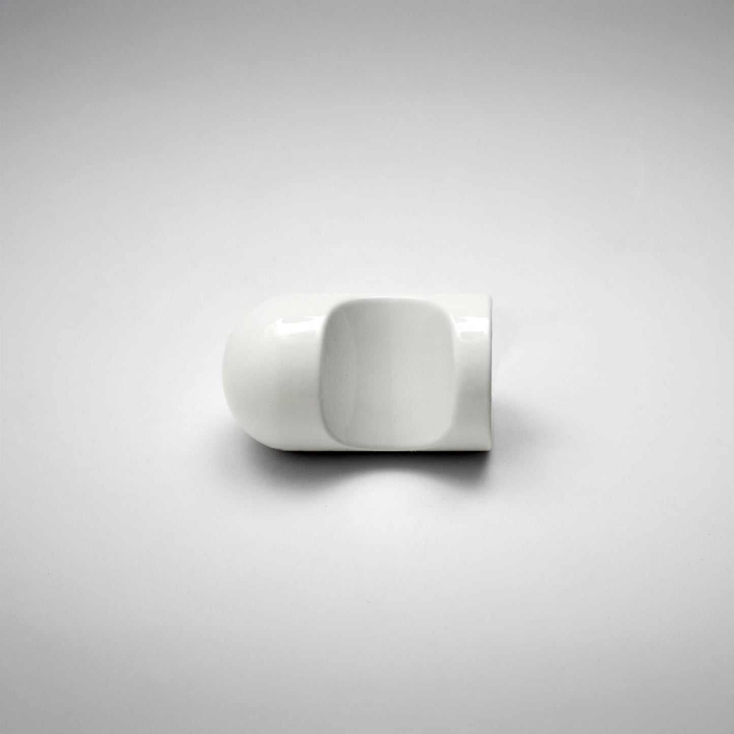 A minimalist white cabinet pull with a unique rounded rectangular shape and a central cutout, shown on a neutral light gray background.